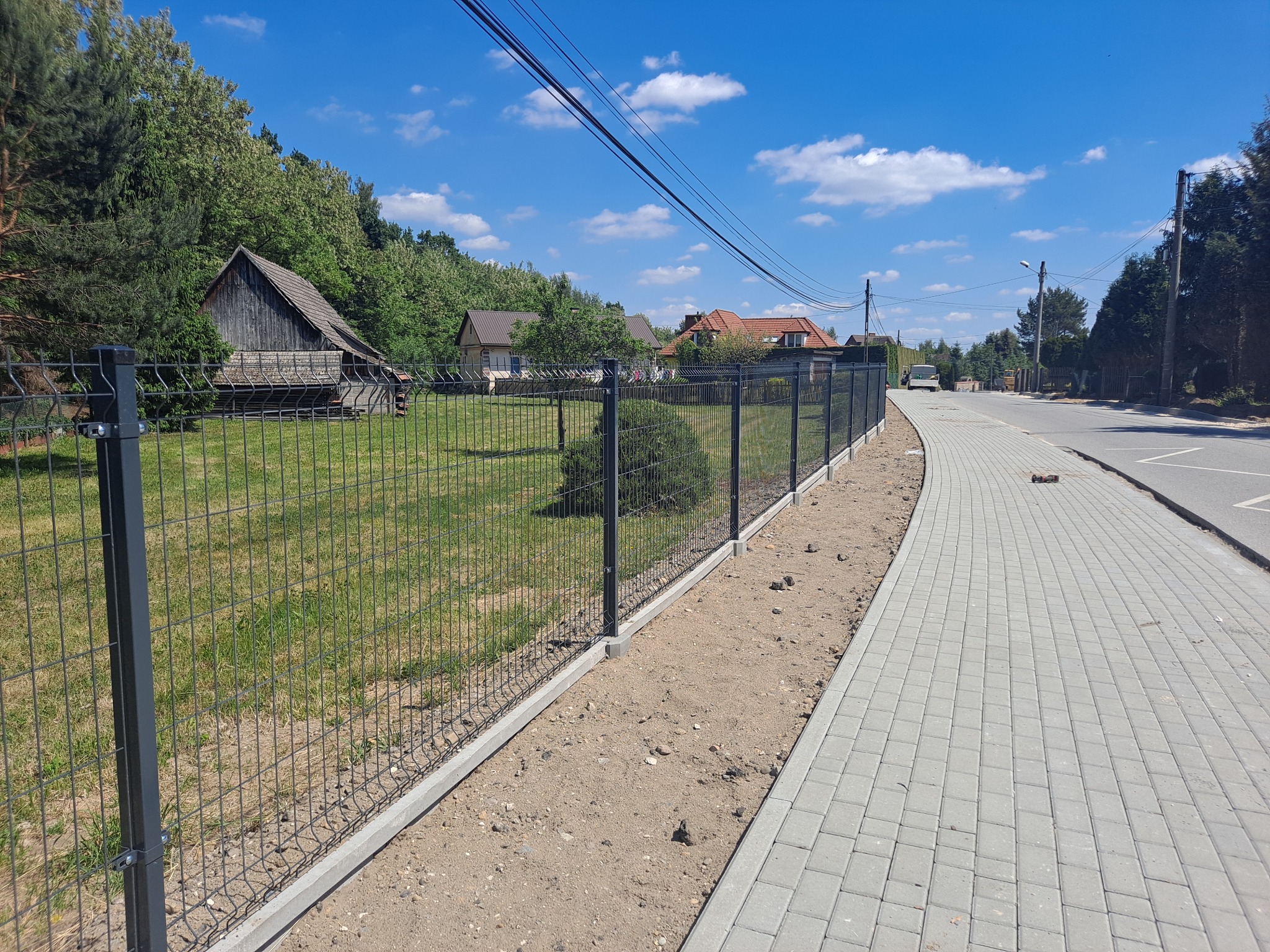 Nowo zamontowane, grafitowe ogrodzenie panelowe z podmurówką betonową, oddzielające posesję od chodnika wyłożonego szarą kostką brukową. W tle wiejska zabudowa i błękitne niebo.