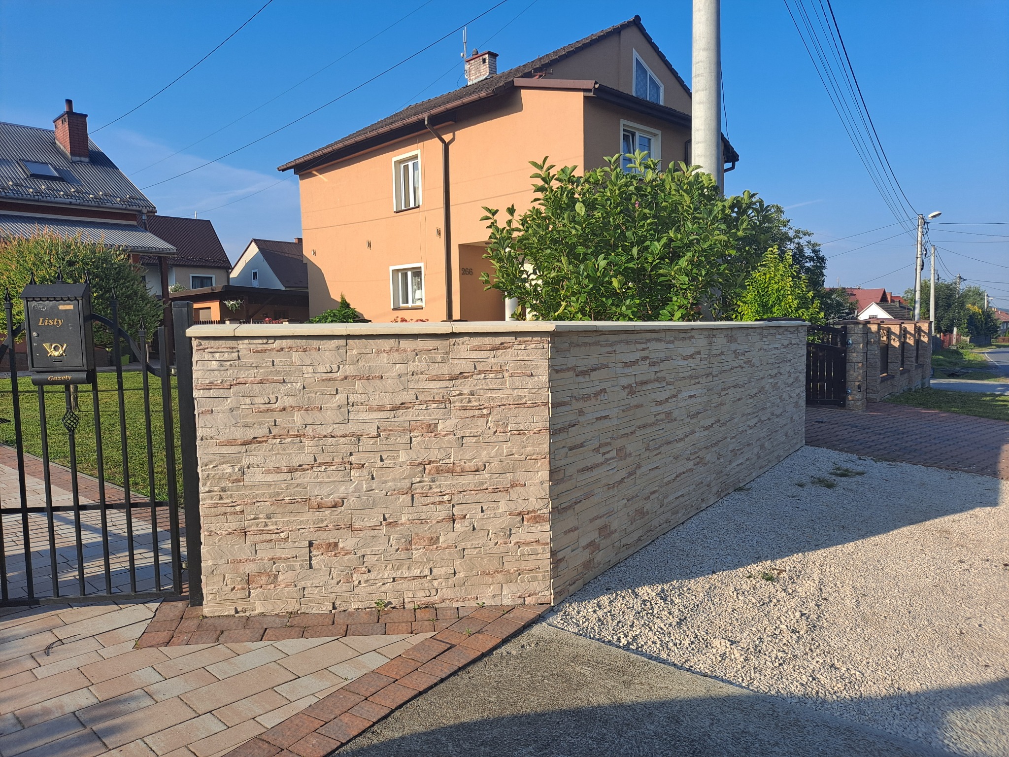 Nowoczesny, niski murek ogrodzeniowy z kamienia elewacyjnego w kolorze beżowym, oddzielający posesję od chodnika; widoczna brama wjazdowa i skrzynka na listy.