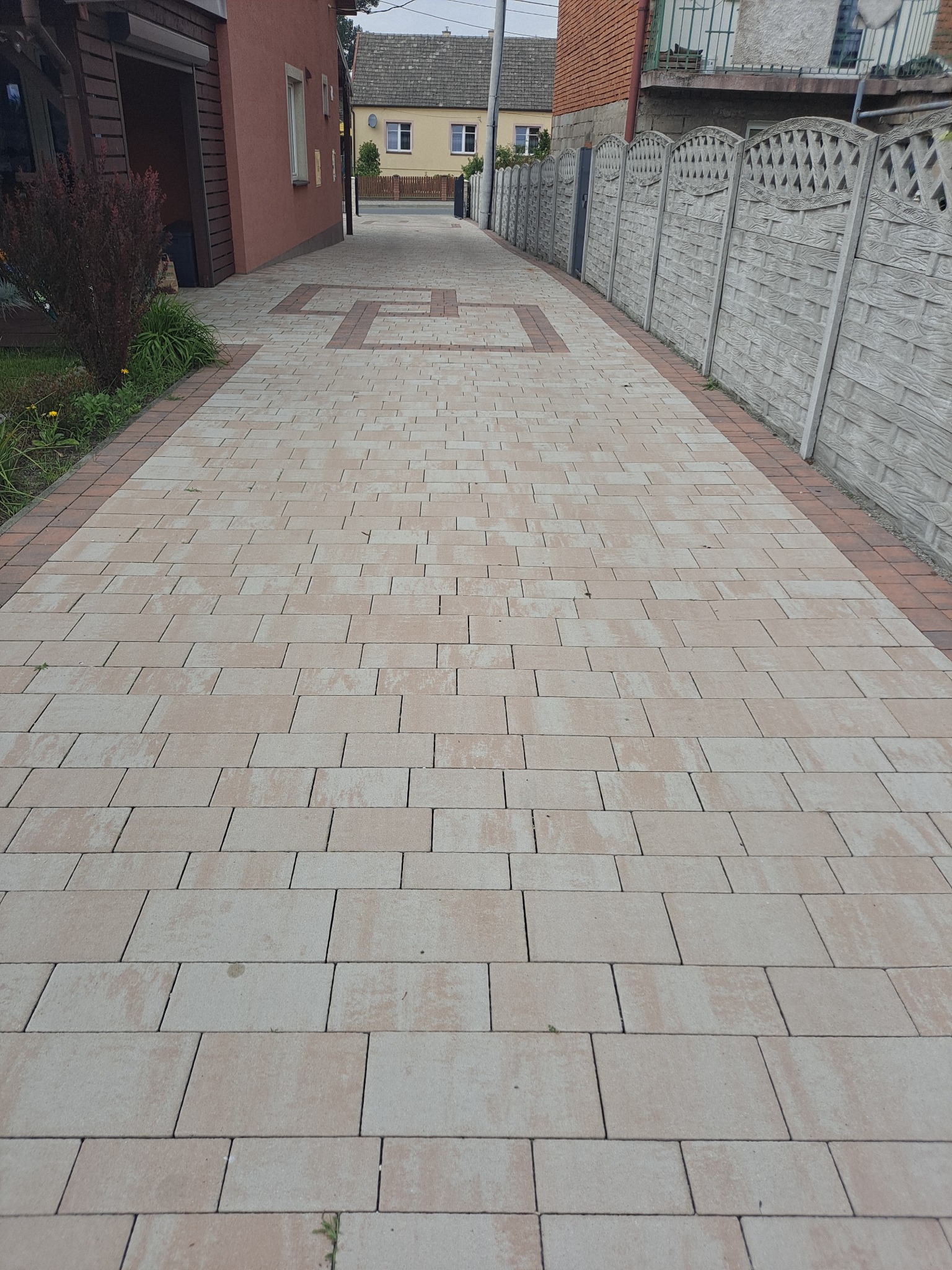 Długa, wybrukowana droga z jasnych prostokątnych kostek z obramowaniem z ceglastoczerwonej kostki, prowadząca między budynkiem mieszkalnym a betonowym ogrodzeniem.