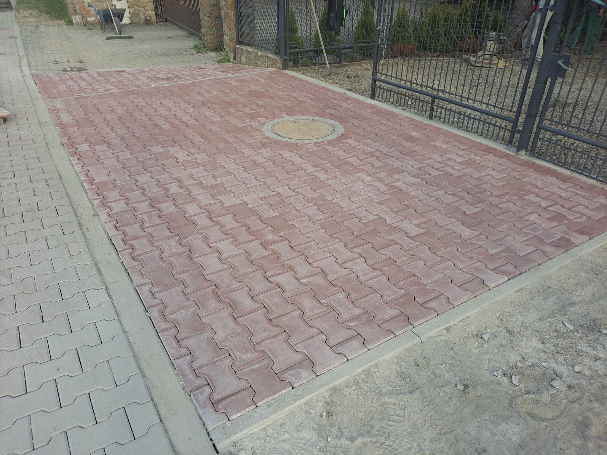 Nowo ułożona, czerwona kostka brukowa w kształcie litery H, z szarą obwódką i okrągłą studzienką kanalizacyjną pośrodku, prowadząca do metalowej bramy w tle.