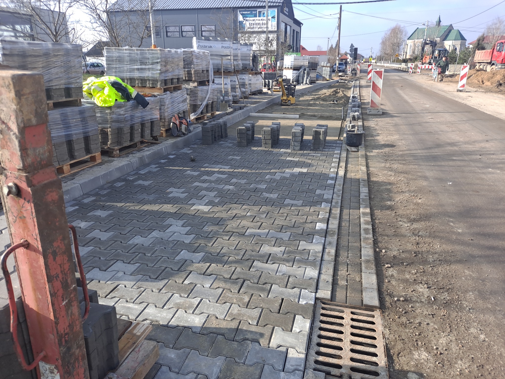 Ułożona kostka brukowa na jezdni, palety z kostką i odzież robocza w tle, widoczne narzędzia i maszyny budowlane, roboty drogowe w trakcie realizacji.