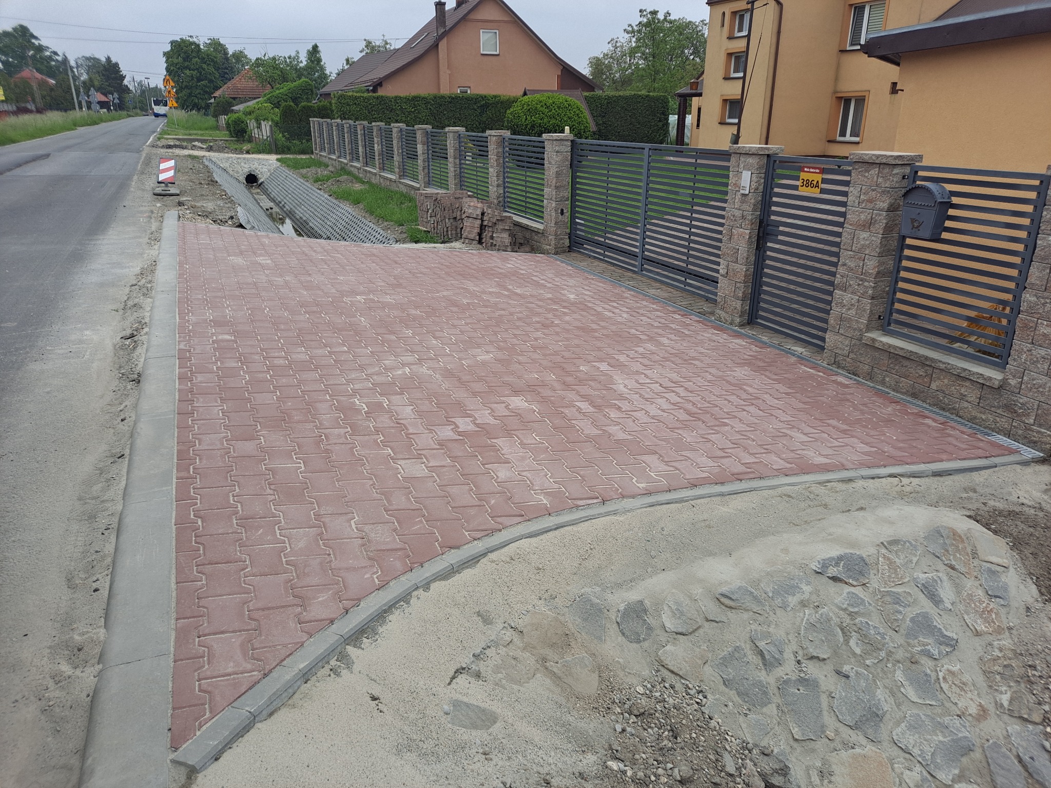 Nowo ułożona, czerwona kostka brukowa na podjeździe, obok asfaltowej drogi i nowoczesnego ogrodzenia z szarej stali z kamiennymi słupkami. Widoczny fragment posesji z budynkami mieszkalnymi w tle.