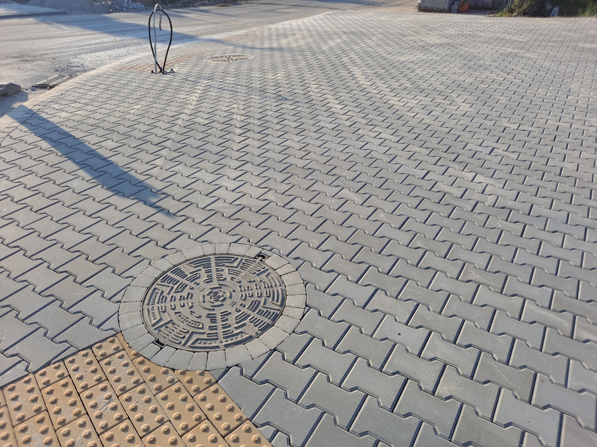 Szara kostka brukowa ułożona w jodełkę, z okrągłą studzienką kanalizacyjną wkomponowaną w nawierzchnię oraz fragmentem żółtej kostki z wypustkami dla osób niedowidzących.