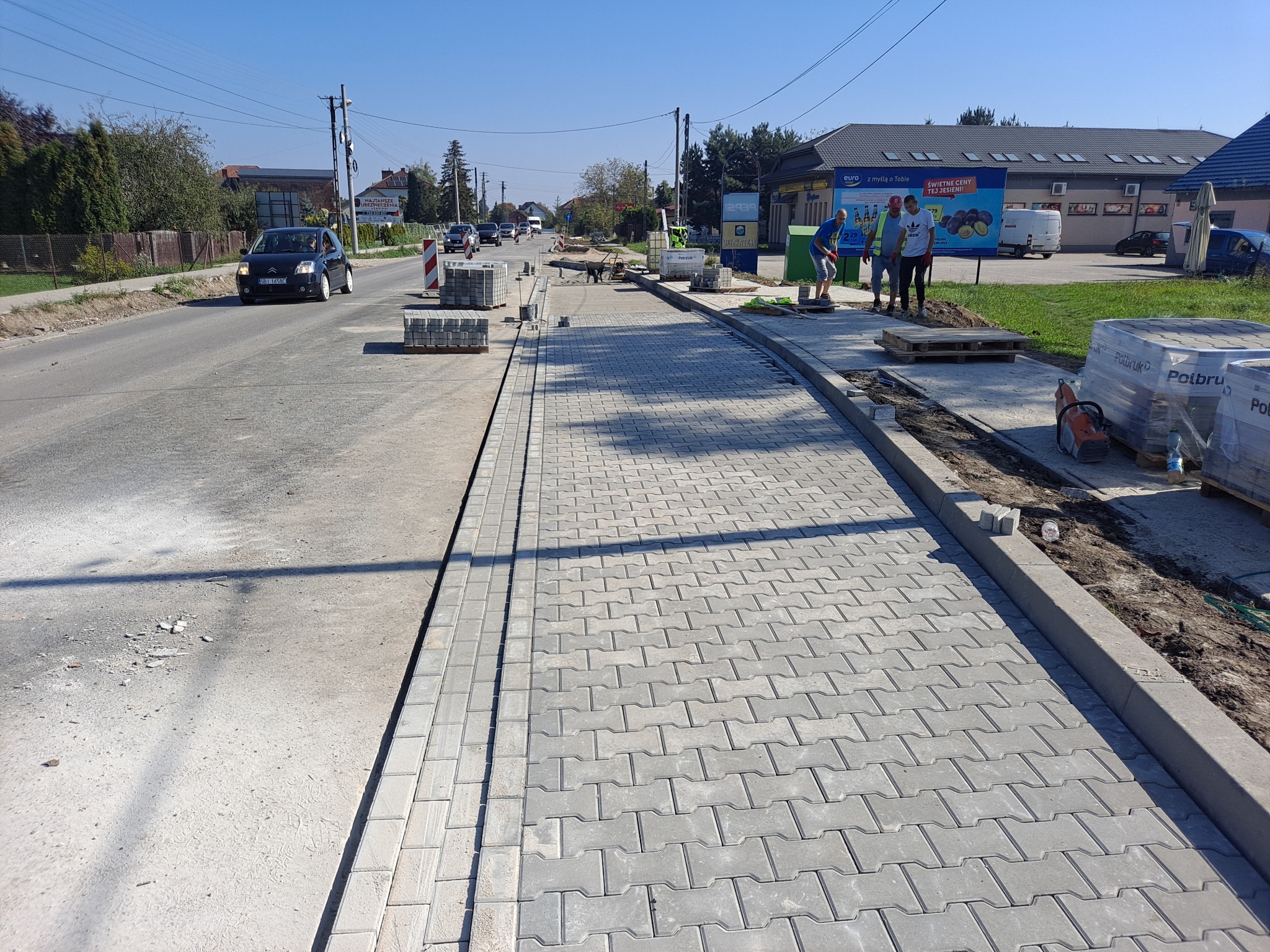 Nowy chodnik z kostki brukowej w Jodłówce, w trakcie układania. Widoczni pracownicy, maszyna do cięcia kostki, palety z kostką i ruch uliczny w tle.