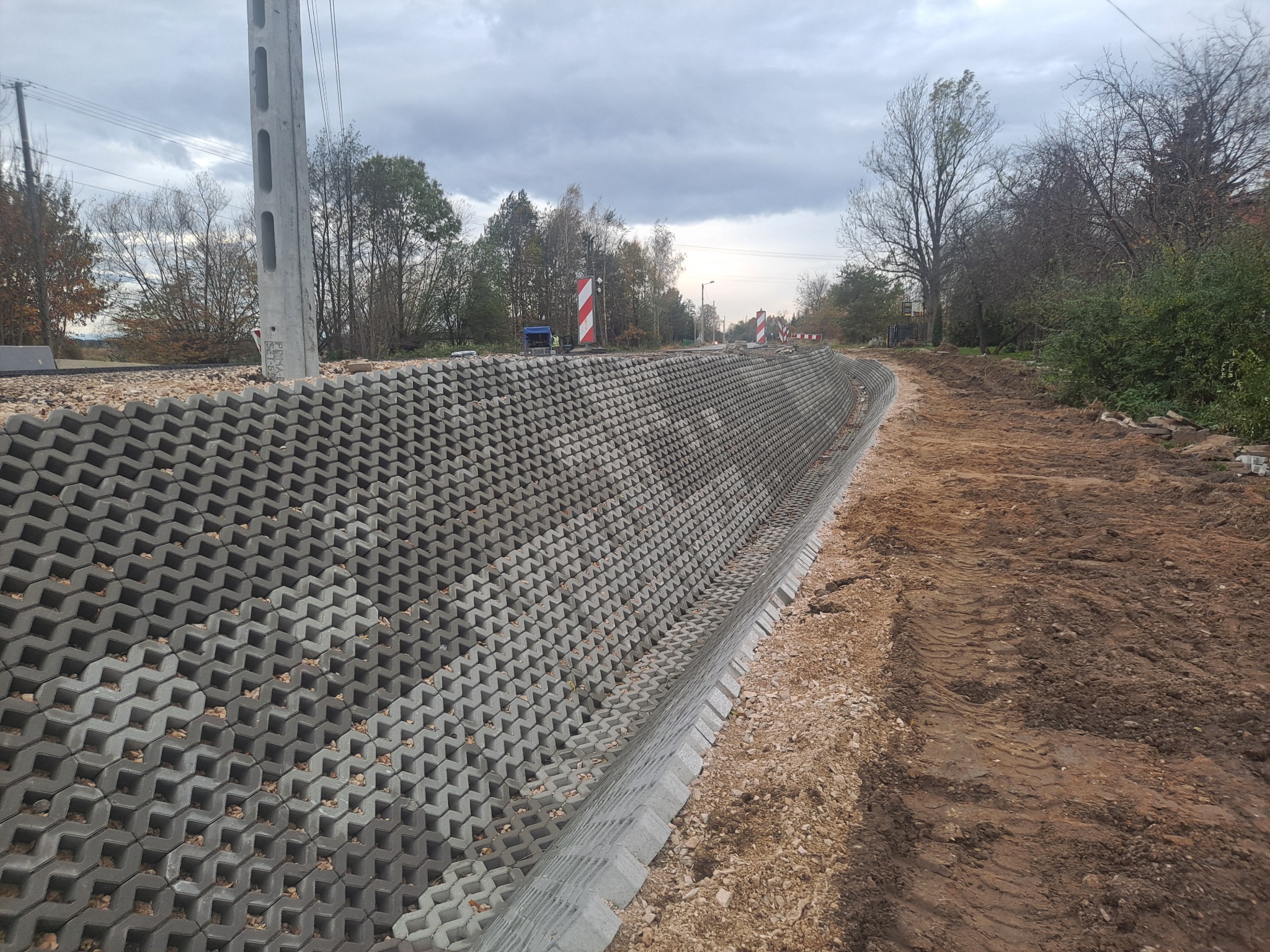 Wzmocnienie skarpy przy drodze za pomocą ażurowych betonowych elementów, widoczne ślady opon i słup energetyczny w tle.