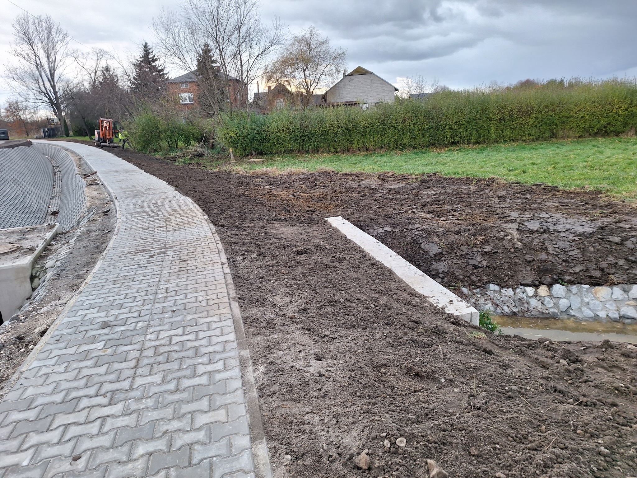 Świeżo ułożona, szara kostka brukowa wzdłuż drogi z widocznym wykopem pod rzekę, z pracującym w tle małym traktorem i robotnikiem w żółtej kamizelce, przy pochmurnym niebie.