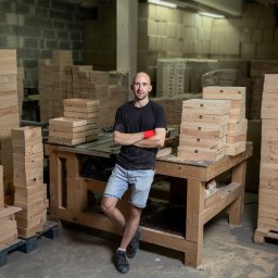grobelnybartek com - Zdjęcie wizerunkowe we Wrocławiu przedstawia mężczyznę z założonymi rękami, ubranego w czarną koszulkę i jeansowe szorty, stojącego w warsztacie stolarskim obok stosów drewnianych pudełek z wieczkami.