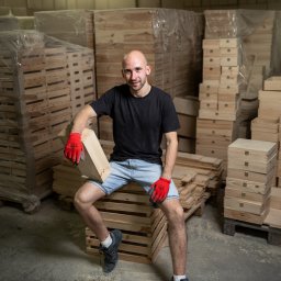 grobelnybartek com - Portret uśmiechniętego mężczyzny w czerwonych rękawicach roboczych, siedzącego na stosie drewnianych skrzynek w magazynie, trzymającego jedną skrzynkę w ręku.