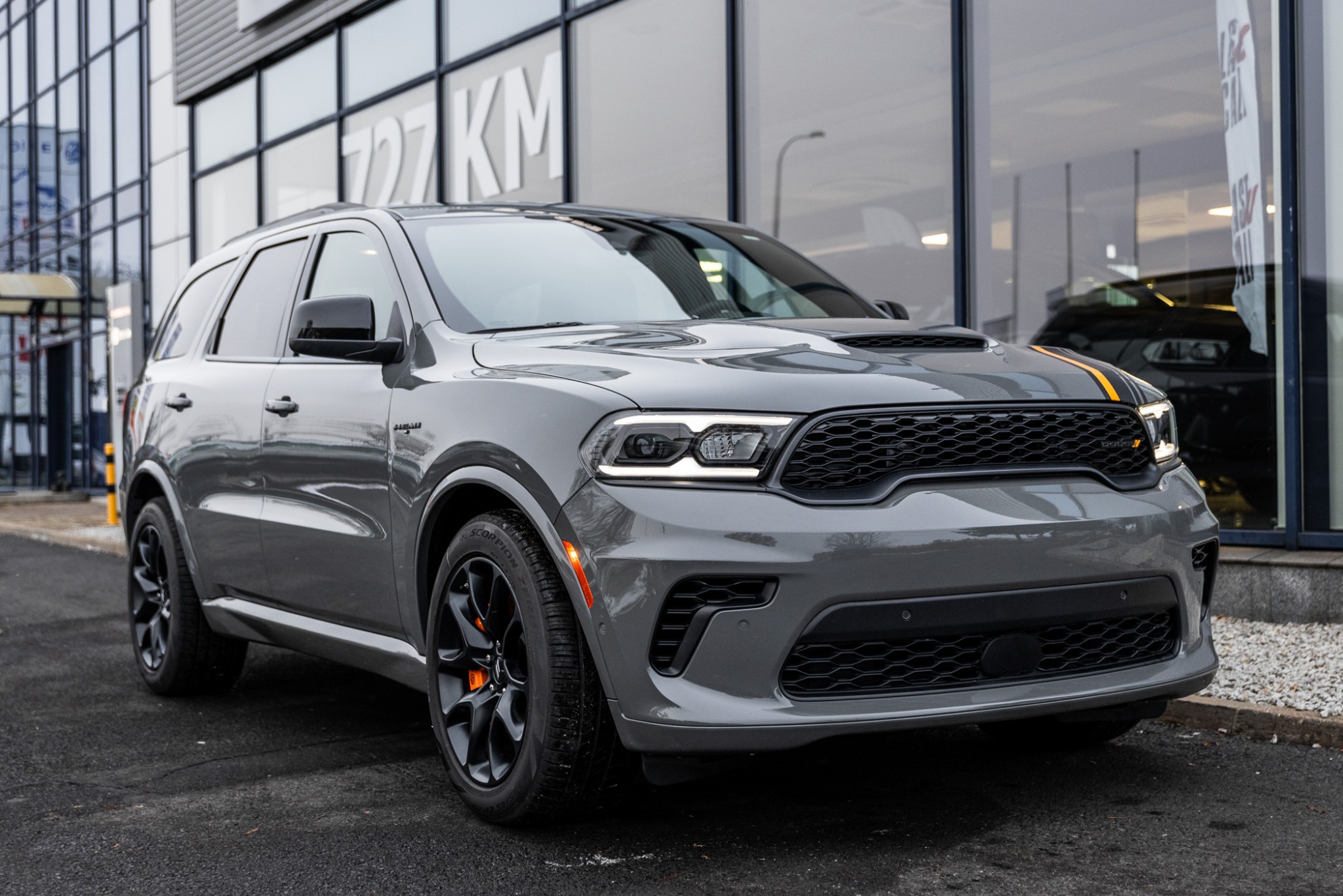 Szary Dodge Durango z czarnymi felgami i pomarańczowymi zaciskami hamulcowymi, stojący przed salonem samochodowym z przeszkloną fasadą.