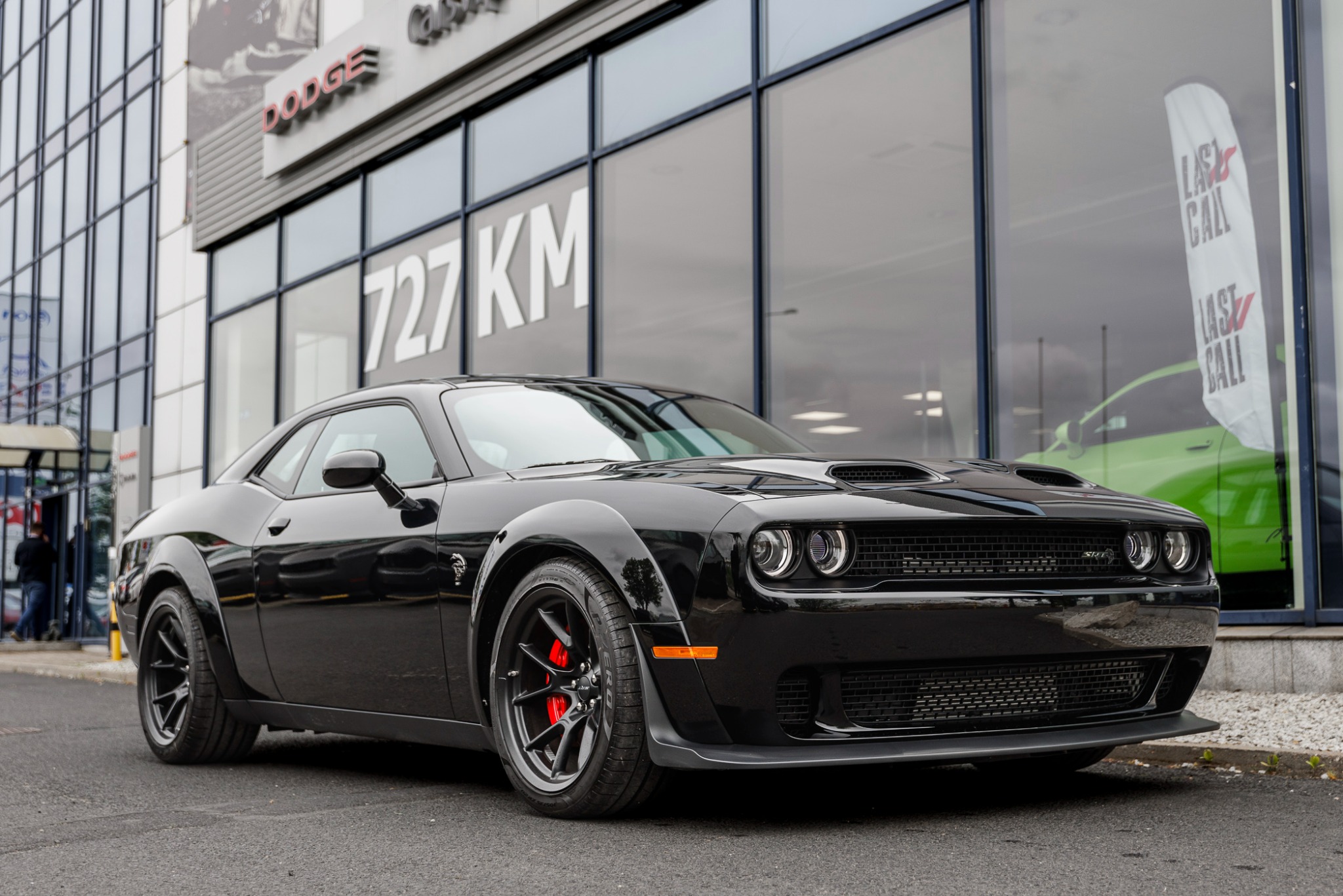 Czarny Dodge Challenger SRT Demon z czerwonymi zaciskami hamulcowymi przed salonem samochodowym z napisem '727 KM' na elewacji.