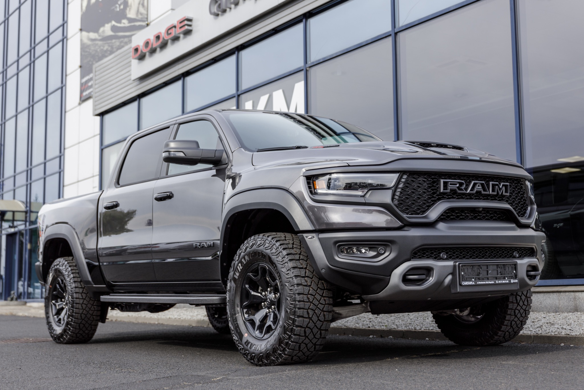 Szary pickup RAM TRX zaparkowany przed nowoczesnym budynkiem z dużymi oknami i logo Dodge na fasadzie.