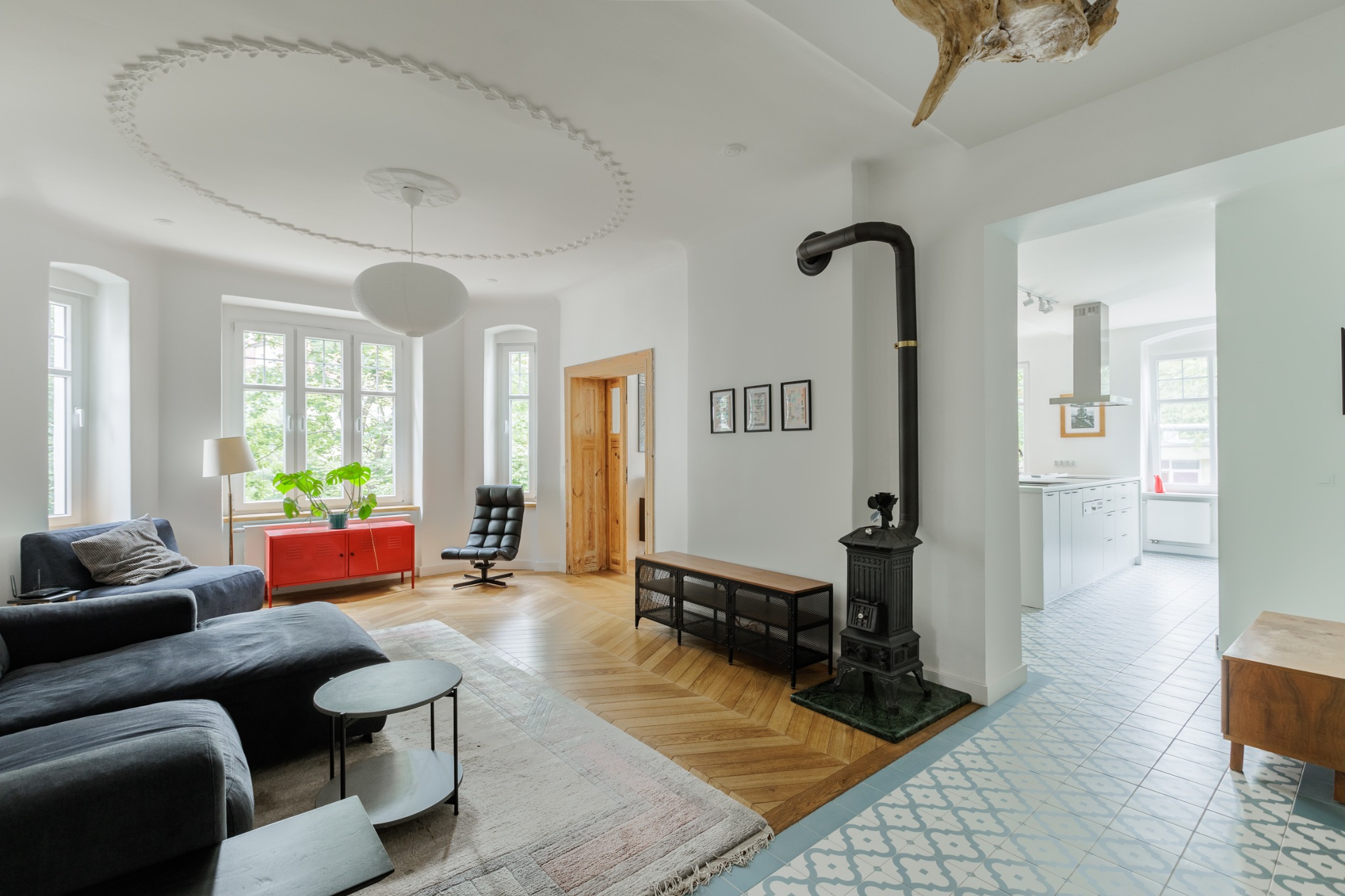 Jasne, przestronne wnętrze salonu we Wrocławiu z parkietem w jodełkę, ciemnoszarą sofą, czerwonym kredensem, czarnym piecem kaflowym i dekoracyjną sztukaterią na suficie.