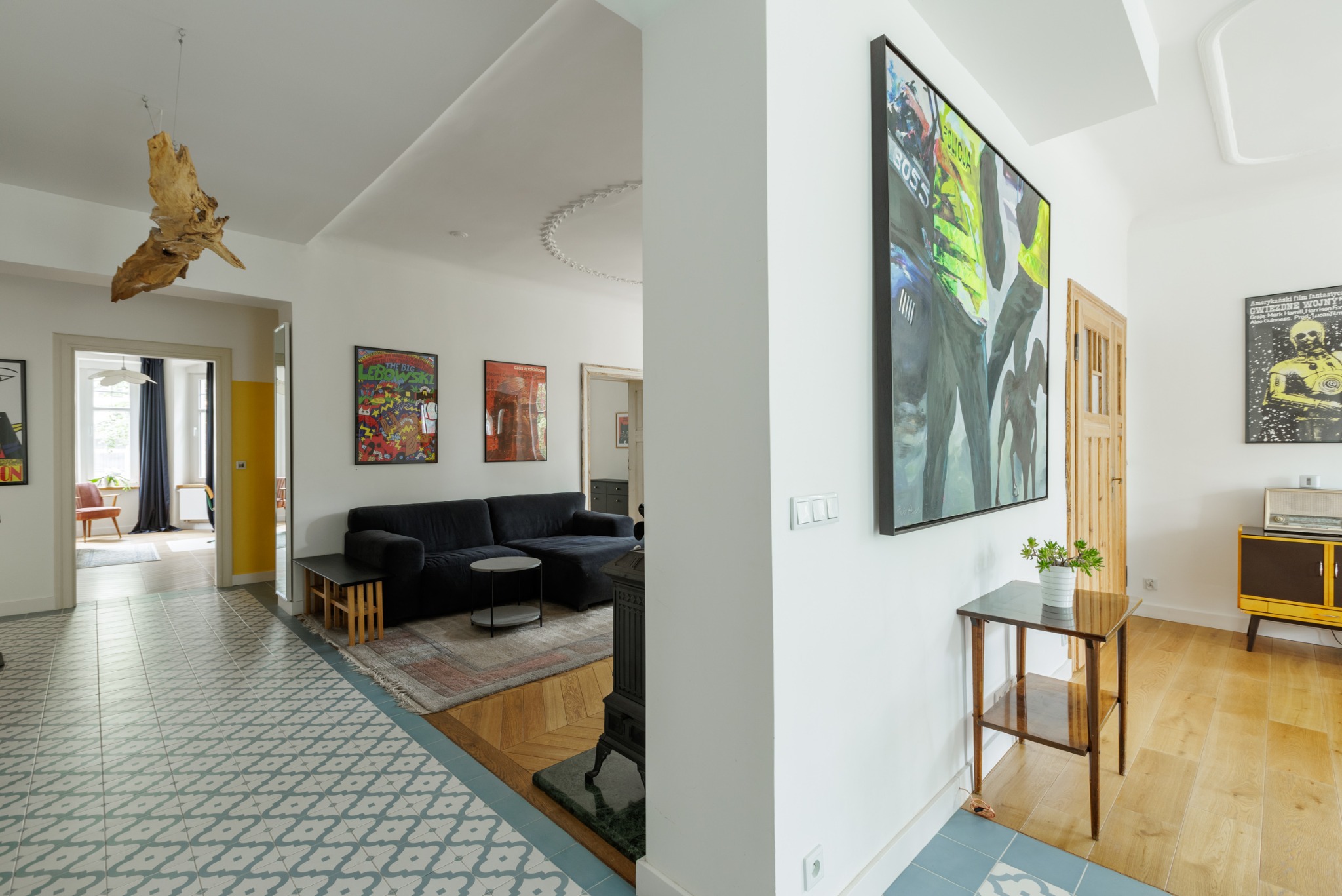 Jasne wnętrze mieszkania z eklektycznym stylem: wzorzysta podłoga, czarna sofa, drewniana rzeźba pod sufitem i nowoczesne obrazy na ścianach.