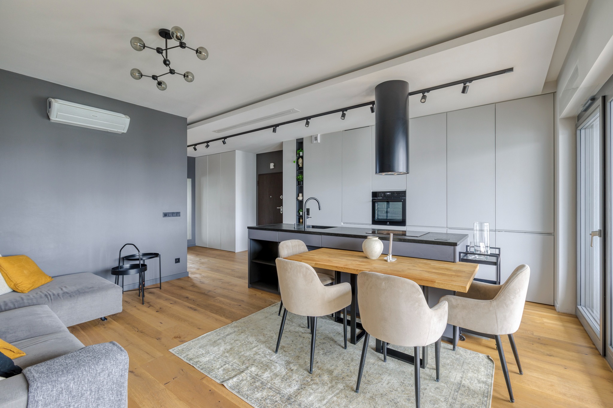 Jasne wnętrze mieszkania z drewnianą podłogą, szarą ścianą i kuchnią z wyspą, oświetlone nowoczesną lampą sufitową i reflektorami, z narożną sofą z żółtymi poduszkami i dywanem pod stołem z krzesłami.