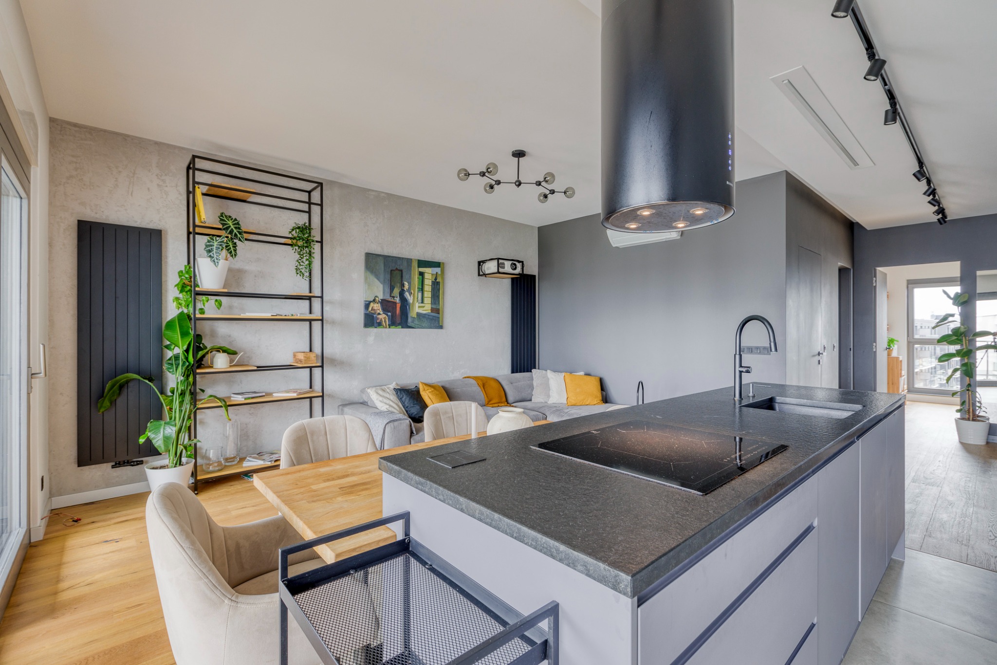 Nowoczesne wnętrze: otwarta kuchnia z wyspą, salon z szarą sofą i żółtymi poduszkami, drewniany stół z krzesłami w odcieniach beżu, czarny okap nad płytą indukcyjną, rośliny doniczkowe na półkach...