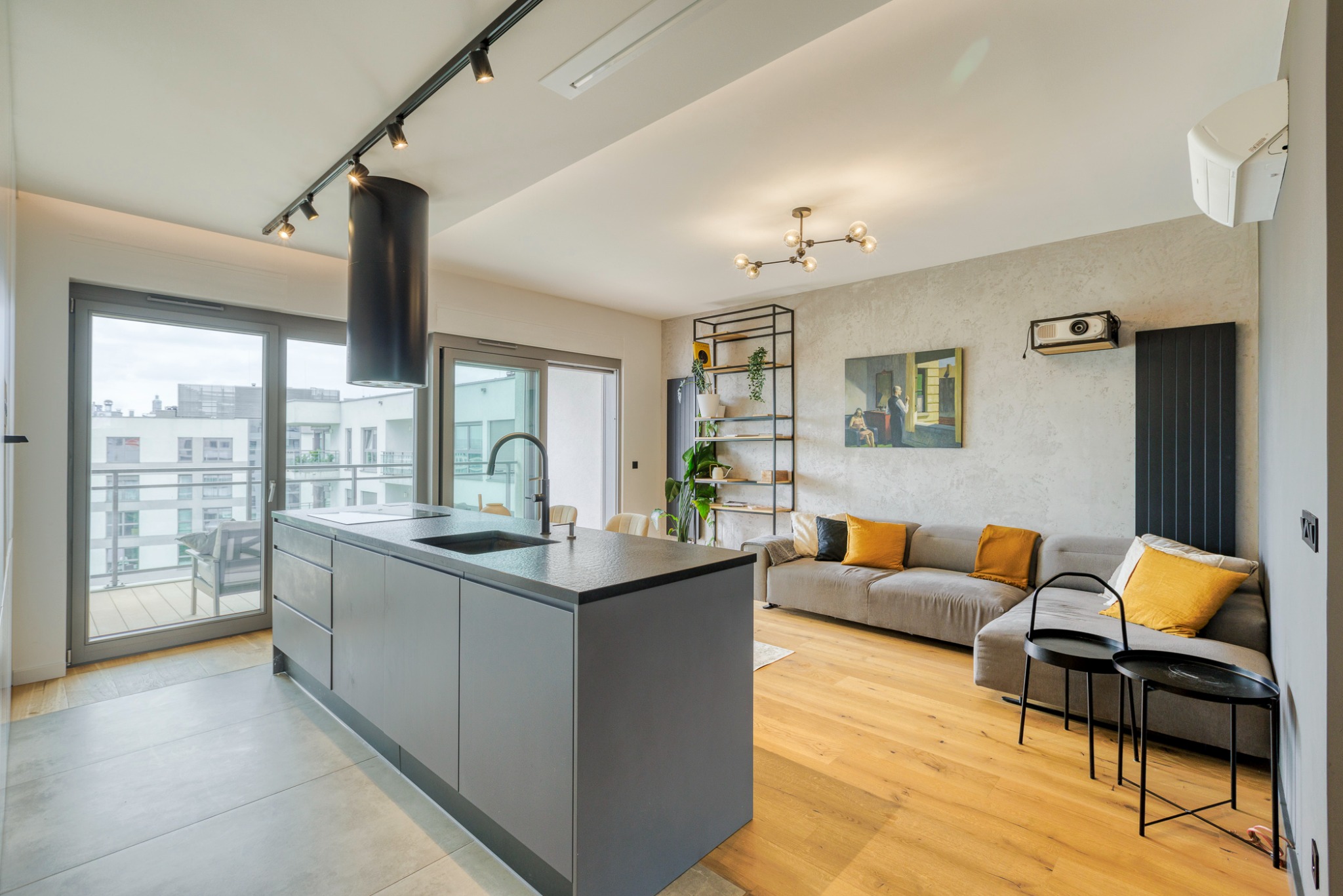 Nowoczesne wnętrze mieszkania z otwartą kuchnią, wyspą kuchenną, szarymi szafkami, drewnianą podłogą i salonem z szarą sofą z żółtymi poduszkami oraz balkonem z widokiem na miasto.