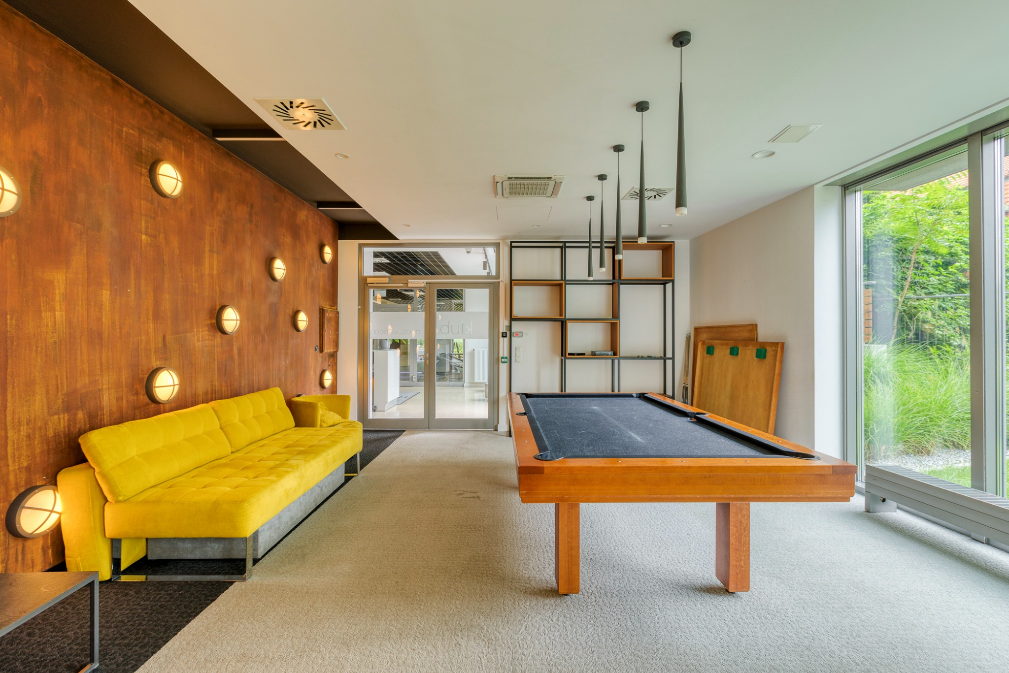 Jasne, przestronne wnętrze z żółtą sofą na tle ściany z oświetleniem punktowym, stołem bilardowym i dużym oknem z widokiem na zieleń.