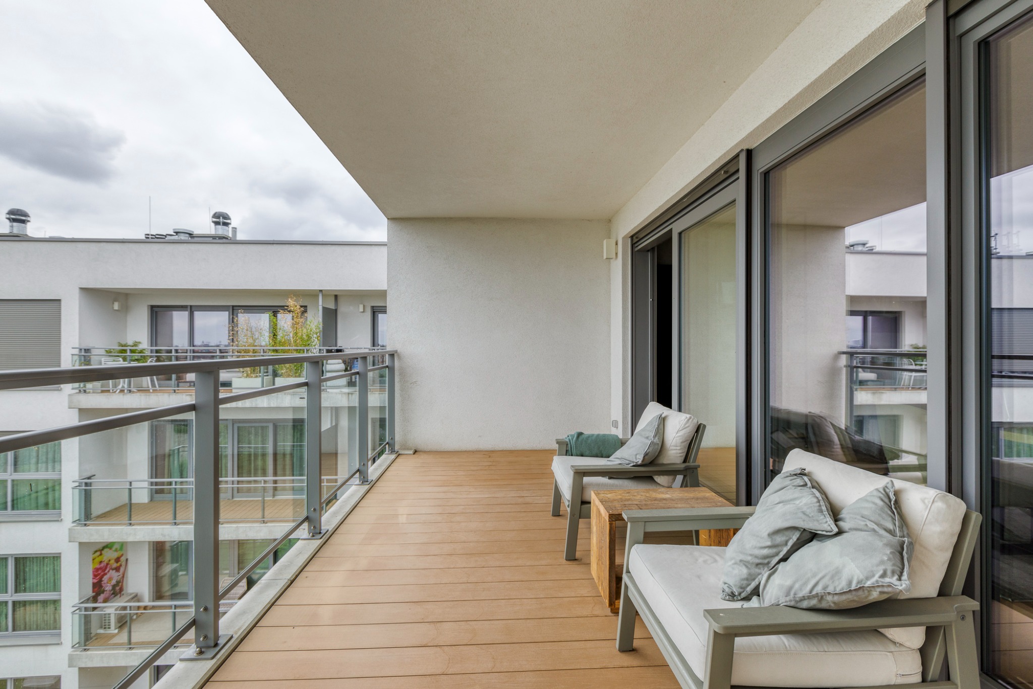 Nowoczesny balkon z drewnianą podłogą, szklaną balustradą i minimalistycznymi meblami wypoczynkowymi w odcieniach szarości. Widok na sąsiedni budynek z balkonami.