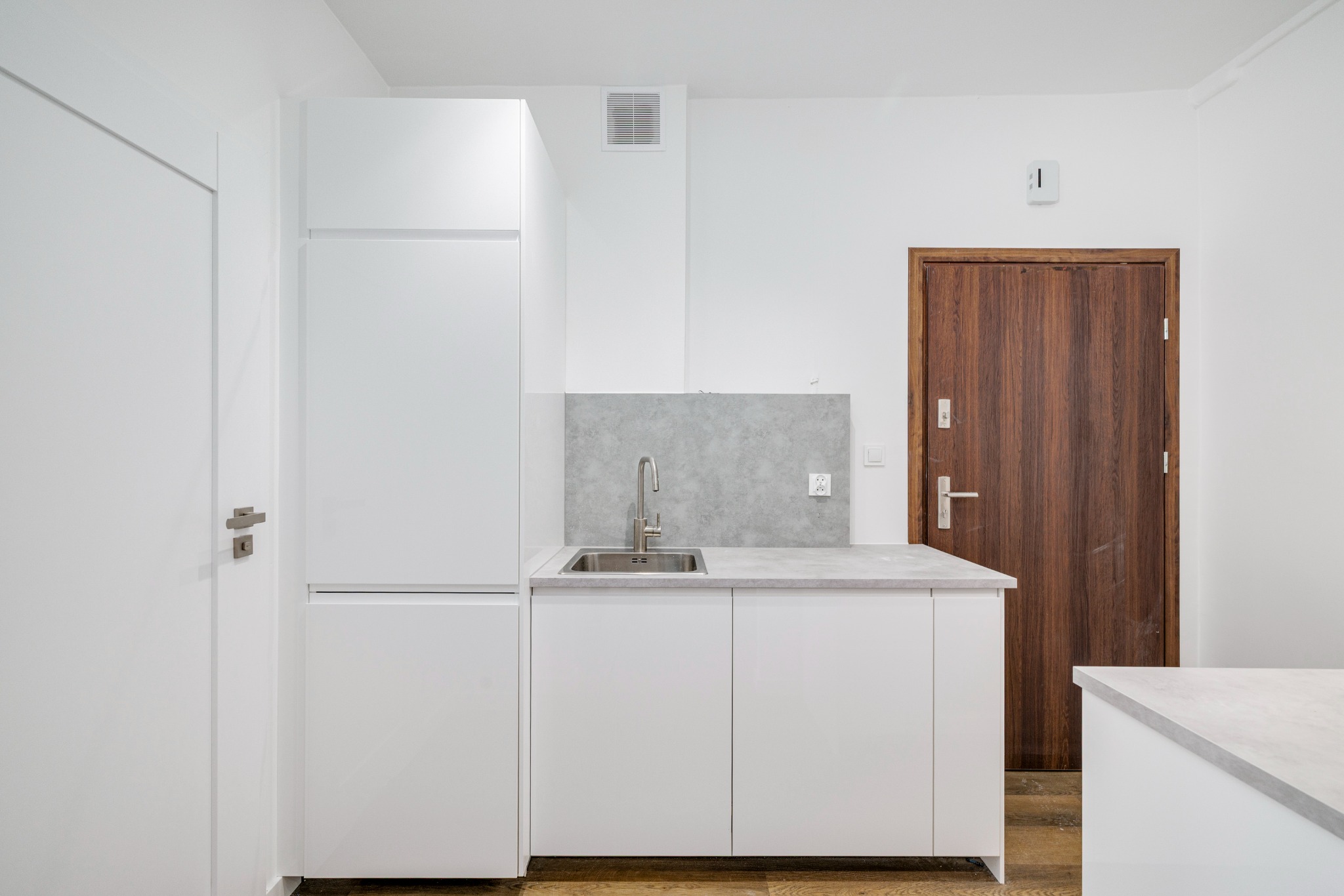 Minimalistyczna, biała zabudowa kuchenna z szarą ścianką nad blatem i drewnianymi drzwiami w tle. Nowoczesny design, proste linie, naturalne światło.