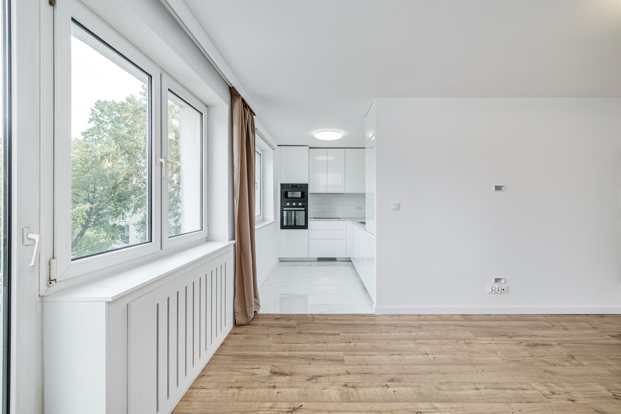 Jasne wnętrze mieszkania z drewnianą podłogą i widokiem na kuchnię. Duże okno z zasłoną wpuszcza światło. Minimalistyczny design, białe ściany.
