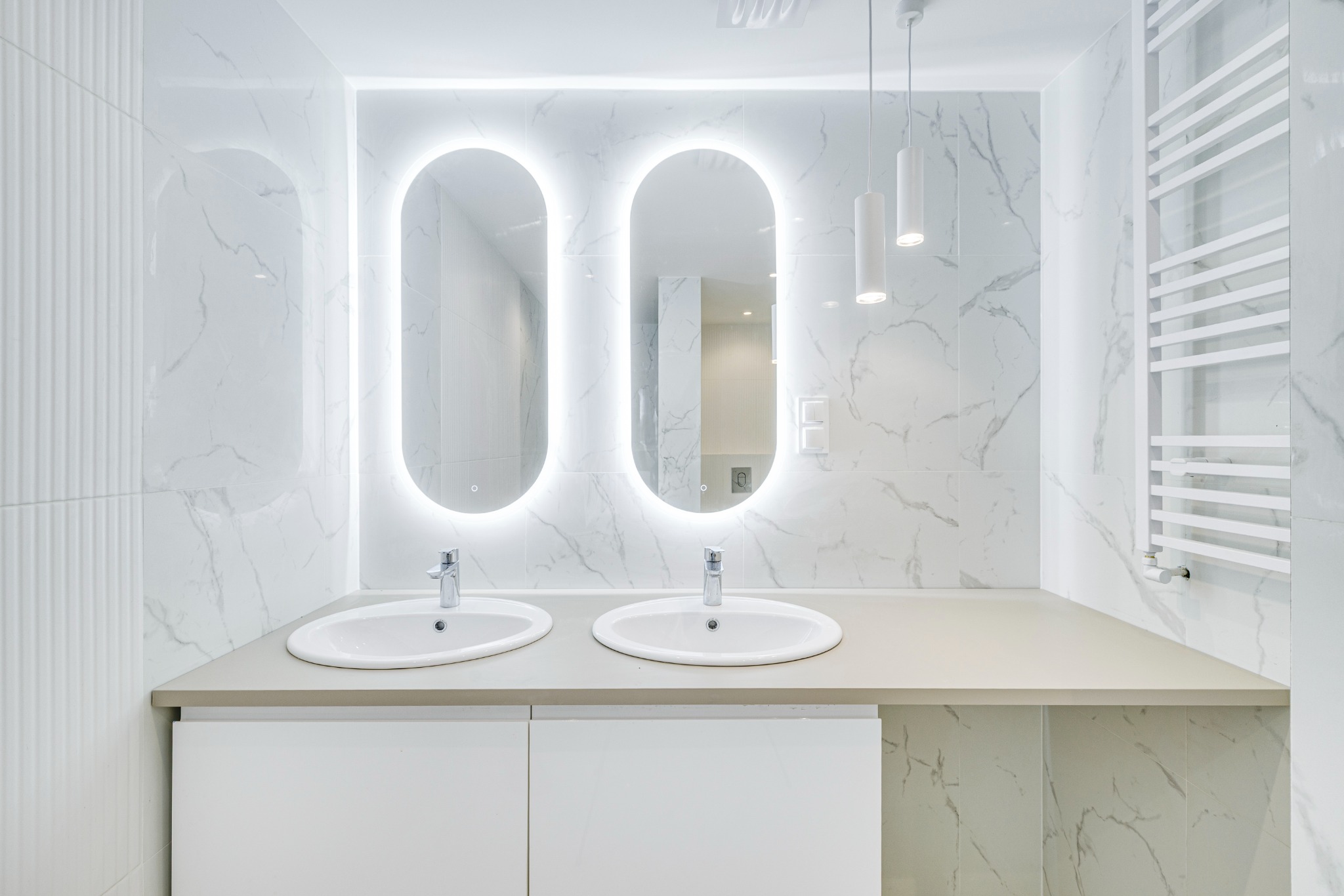 Nowoczesna łazienka w bieli: dwie owalne umywalki z podświetlanymi lustrami na tle marmurowych płytek i grzejnika drabinkowego. Minimalistyczne oświetlenie.