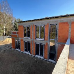 montujeMY - Surowy stan budynku z zamontowanymi oknami PCV w czarnych ramach. Widok z góry na dwukondygnacyjny budynek w trakcie budowy, z oknami z lustrzanym odbiciem.