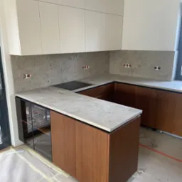 Nowoczesna kuchnia w trakcie wykańczania, z szarym blatem i brązowymi szafkami, widoczne gniazdka elektryczne i zabezpieczona podłoga.