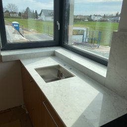 STEC STONE Józef Stec - Biały blat z kamienia z wbudowanym zlewem, widok na okno z zabudową i zewnętrzny krajobraz z budynkami w tle.