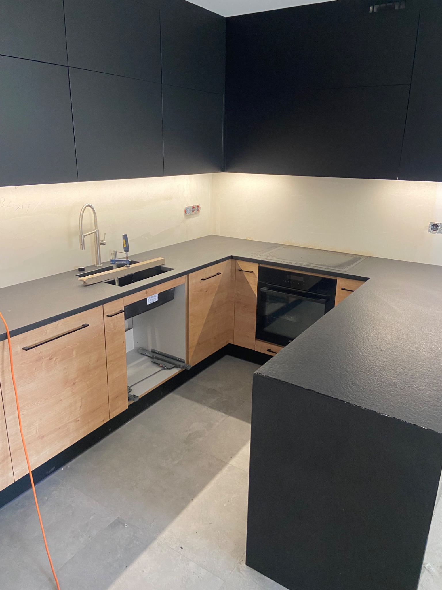 Montaż blatu w kuchni: szary blat roboczy z zamontowanym zlewem i baterią, szafki dolne z drewnopodobnymi frontami, czarne szafki górne, widoczne szyny szuflady, oświetlenie LED pod szafkami.