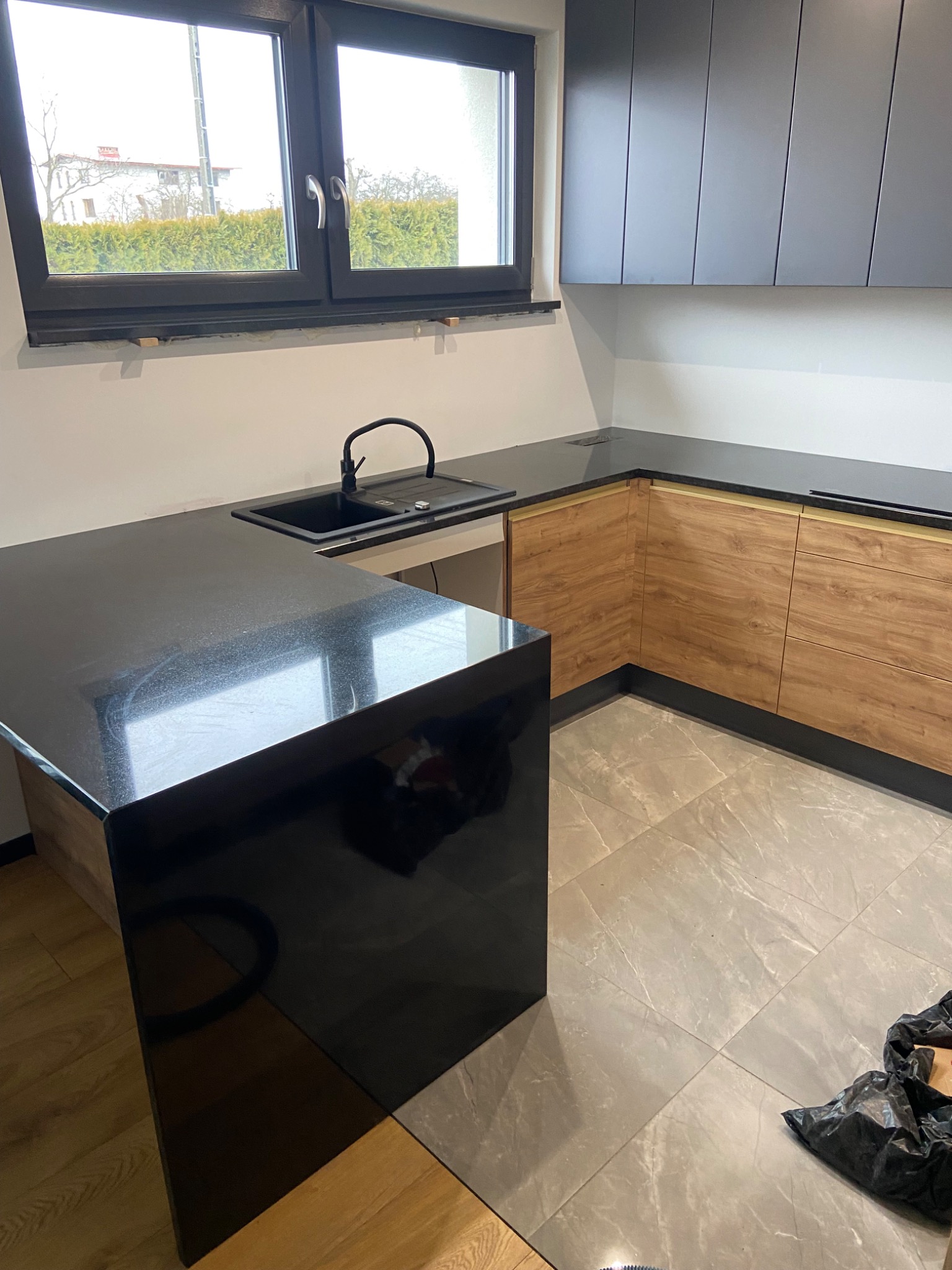 Nowoczesna kuchnia z czarnym blatem i zlewozmywakiem, drewniane szafki dolne i szare górne, widok na okno i fragment podłogi z workiem śmieci w rogu.