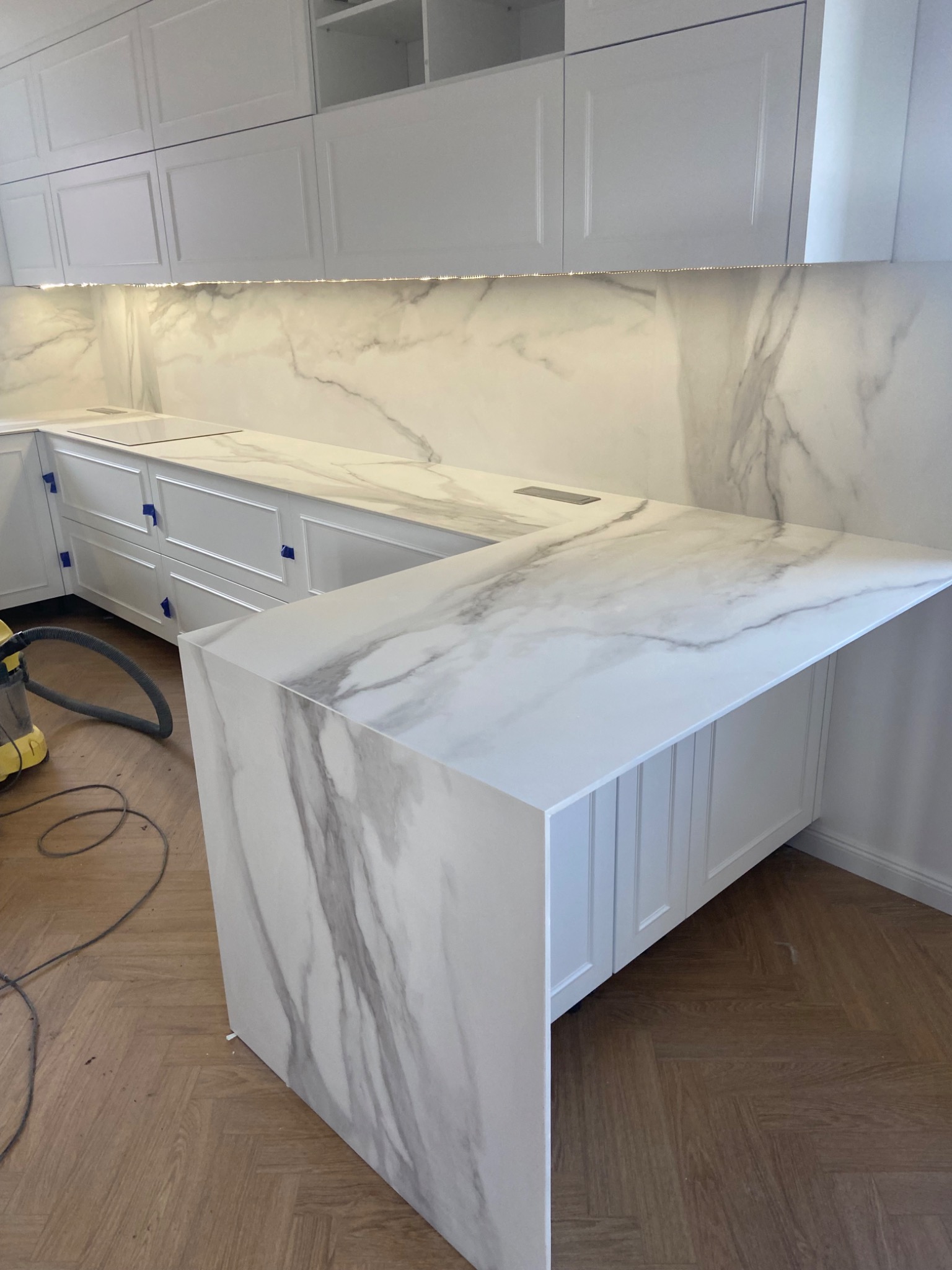 Biała kuchnia z blatem o wzorze marmuru, widoczny fragment odkurzacza na parkiecie w jodełkę.