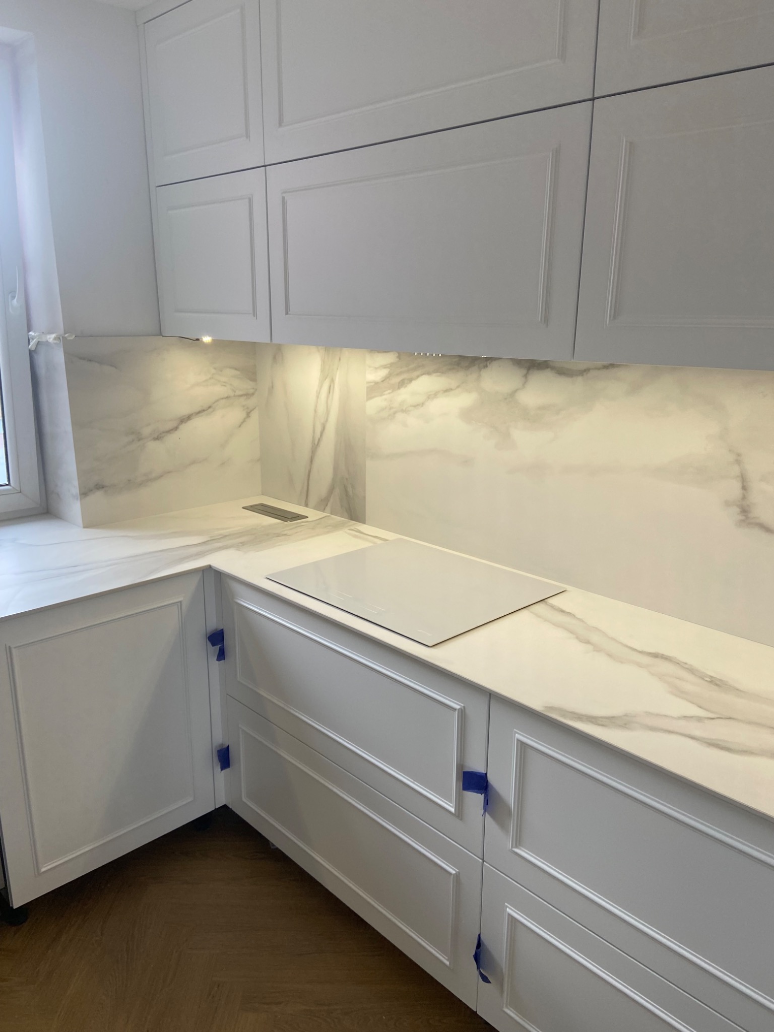 Biała kuchnia z blatem kamiennym imitującym marmur, widoczne zabezpieczenia taśmą malarską na szafkach, płyta indukcyjna i odpływ blatu.