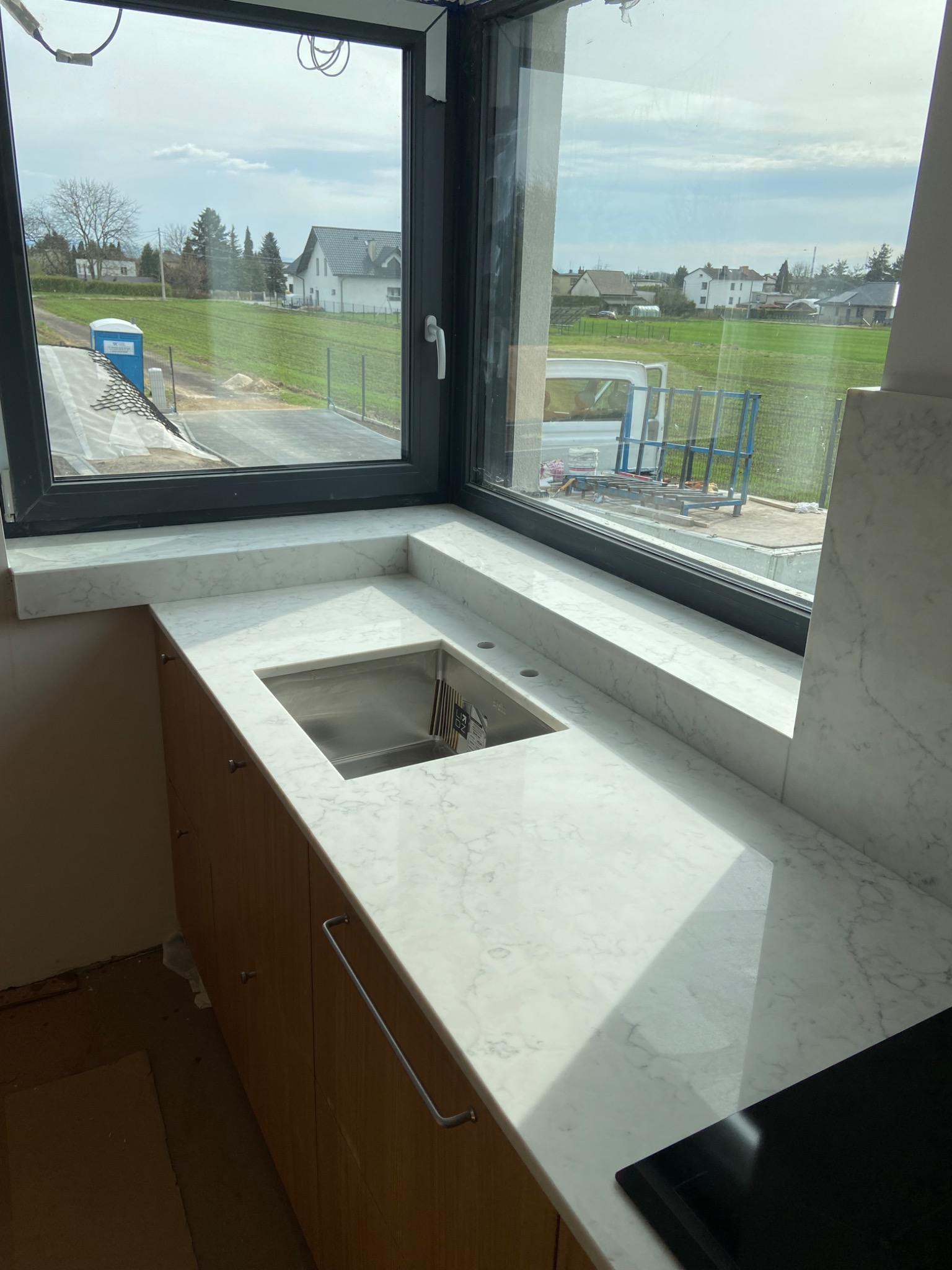 Biały blat z kamienia z wbudowanym zlewem, widok na okno z zabudową i zewnętrzny krajobraz z budynkami w tle.