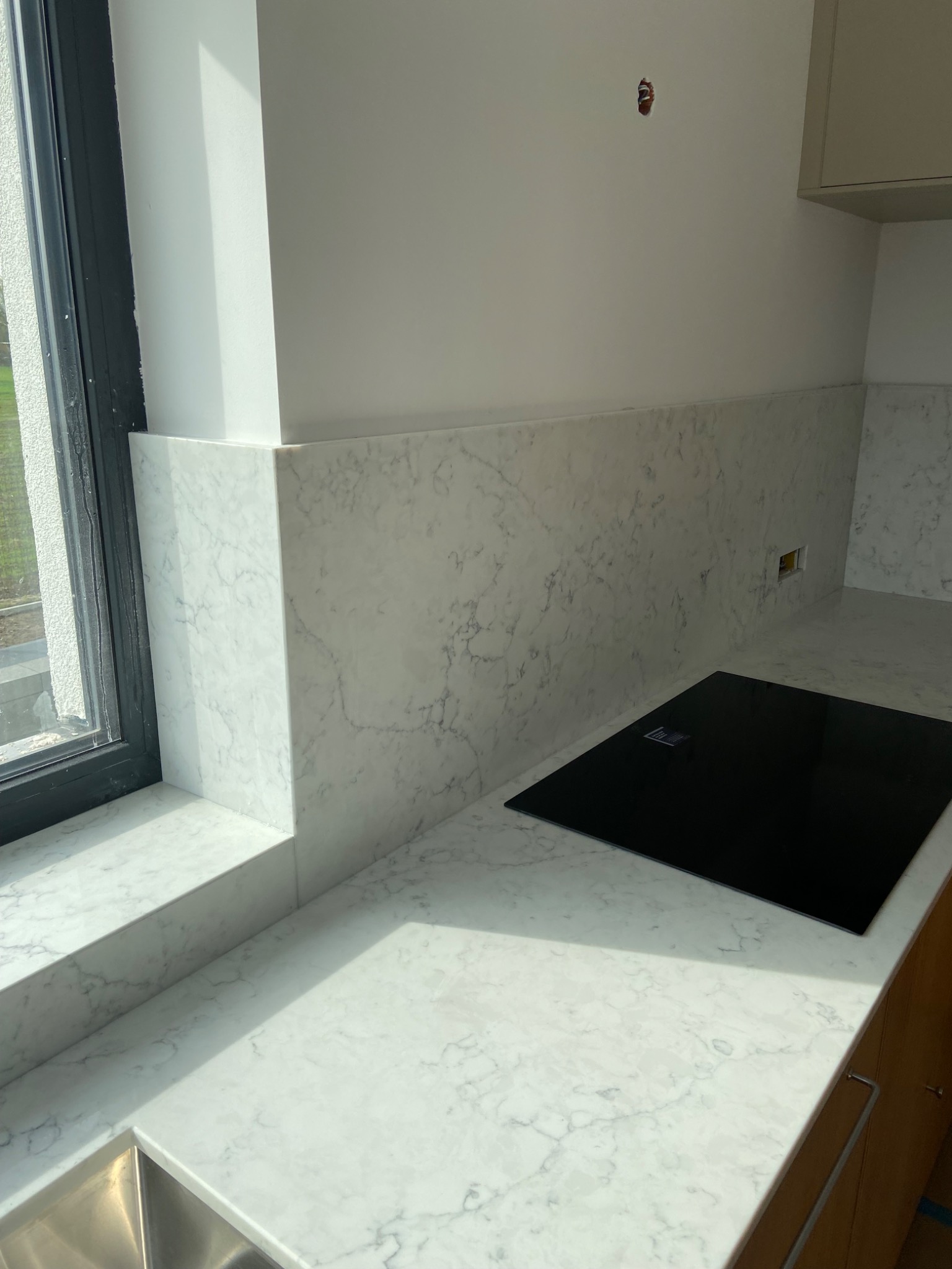 Jasna kuchnia z blatem i panelem ściennym z białego kamienia z wyraźnym marmurkowym wzorem, czarna płyta indukcyjna, widoczny fragment zlewu i okno z czarną ramą.