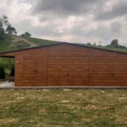 Brązowy garaż blaszany z dwiema bramami i drzwiami bocznymi, zadaszenie po lewej stronie, na betonowej wylewce, trawnik przed garażem, pagórkowaty teren w tle.