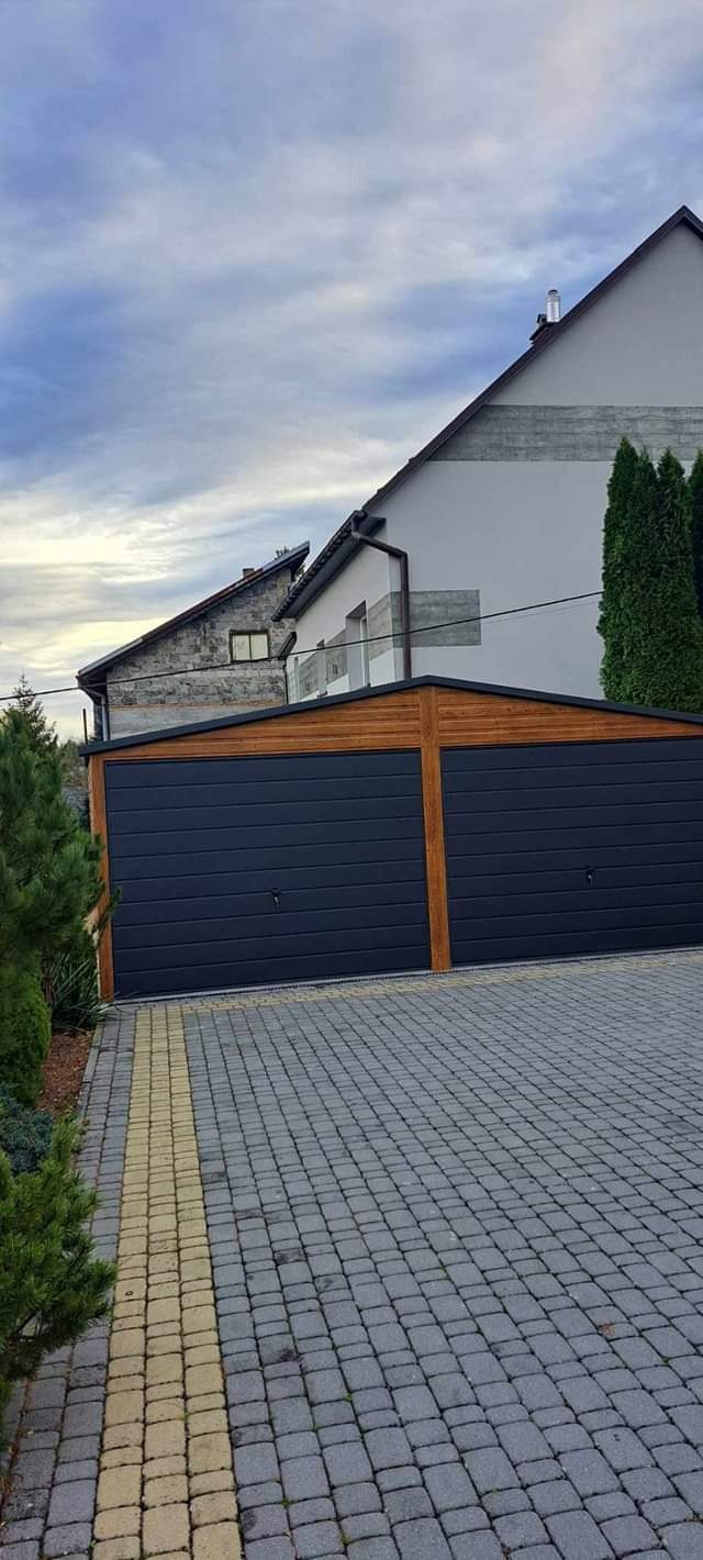 Podwójny garaż blaszany z bramami w kolorze grafitowym, wykończony drewnianą ramą, na posesji z kostki brukowej.