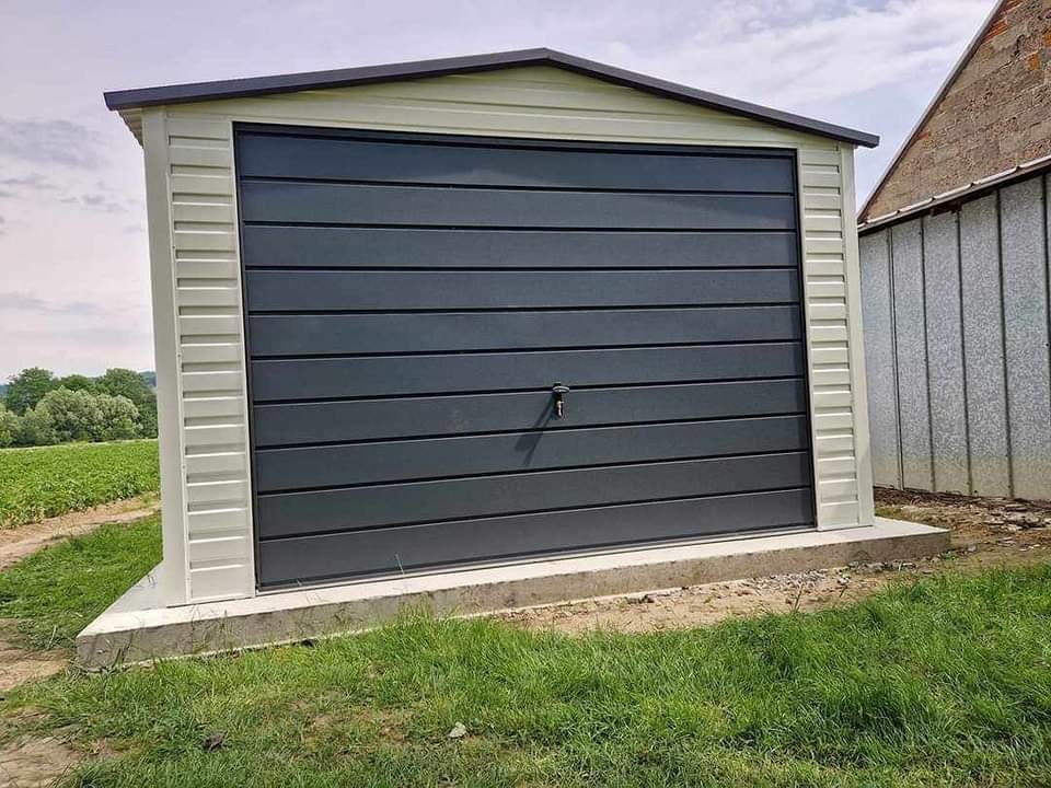 Gotowy, szary garaż z blachy trapezowej z poziomymi przetłoczeniami na betonowej wylewce, widok z zewnątrz na tle zieleni i budynku gospodarczego.