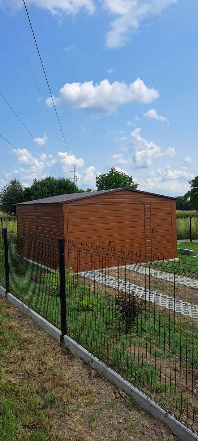Brązowy, metalowy garaż z drzwiami, widziany z zewnątrz, z zielonym trawnikiem, ogrodzeniem z siatki i ścieżką z płyt betonowych, pod błękitnym niebem z chmurami.