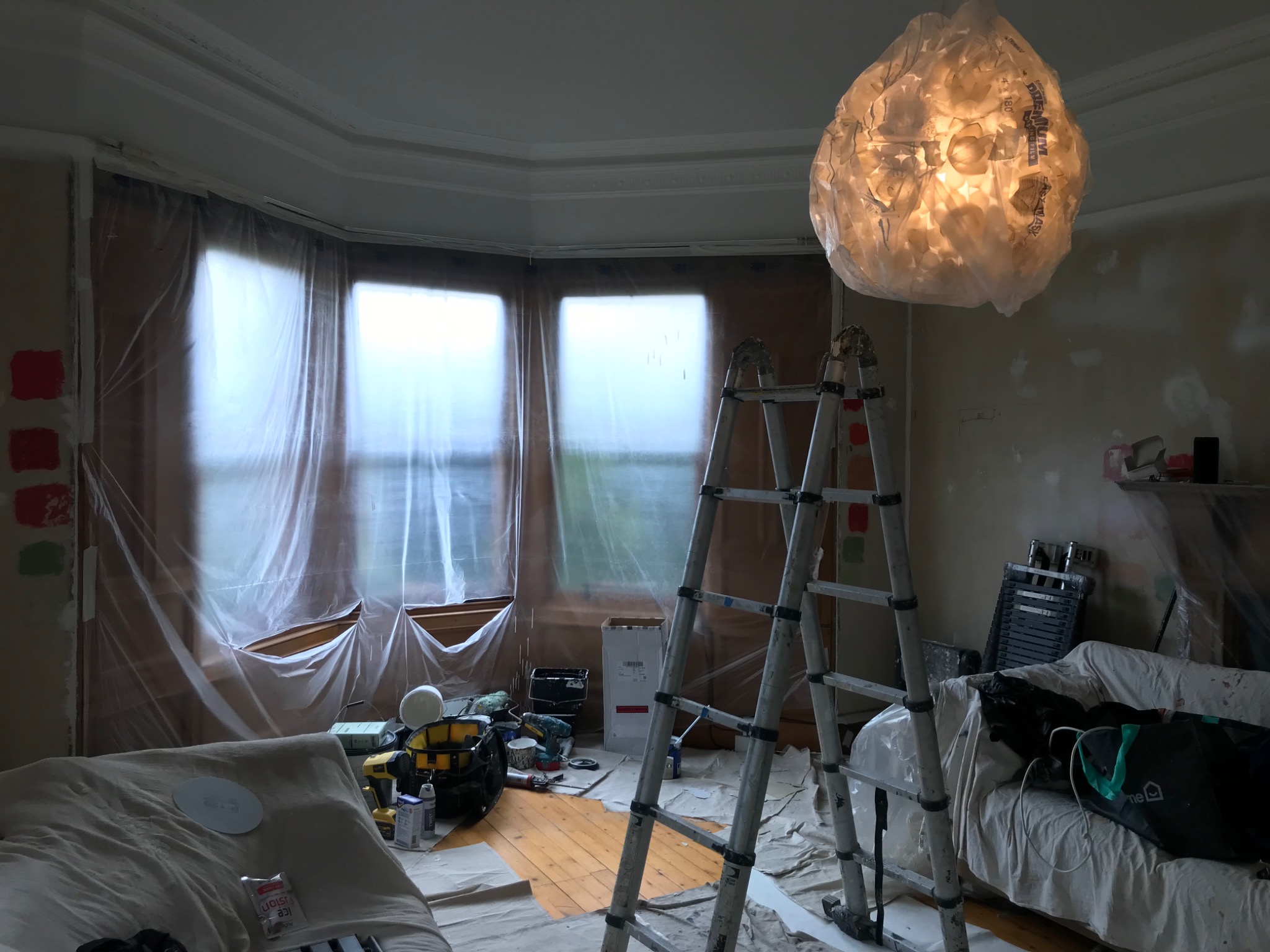 Przygotowanie do malowania pokoju: okna i meble zabezpieczone folią, drabina rozstawiona, widoczne próbniki kolorów na ścianie, nietypowa lampa z tworzywa sztucznego.