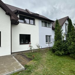 Elewację Jakub Mazowieckie - Dwukondygnacyjny dom z jasną elewacją, brązowym dachem i oknami. Część elewacji w odcieniu szarości. W tle pochmurne niebo i zieleń. Widok z poziomu gruntu.