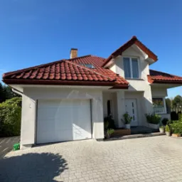 Biały dom jednorodzinny z czerwoną dachówką, garażem i brukowanym podjazdem w słoneczny dzień. Widok z poziomu gruntu, podkreślający estetykę elewacji.