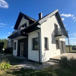 Dwukondygnacyjny dom z balkonami, wykończony w bieli i szarości, na tle błękitnego nieba. Widoczne elementy architektury i otoczenie domu.