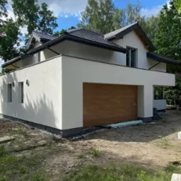 Nowoczesny, dwukondygnacyjny dom z jasną elewacją i brązową bramą garażową, otoczony zielenią i błękitnym niebem.