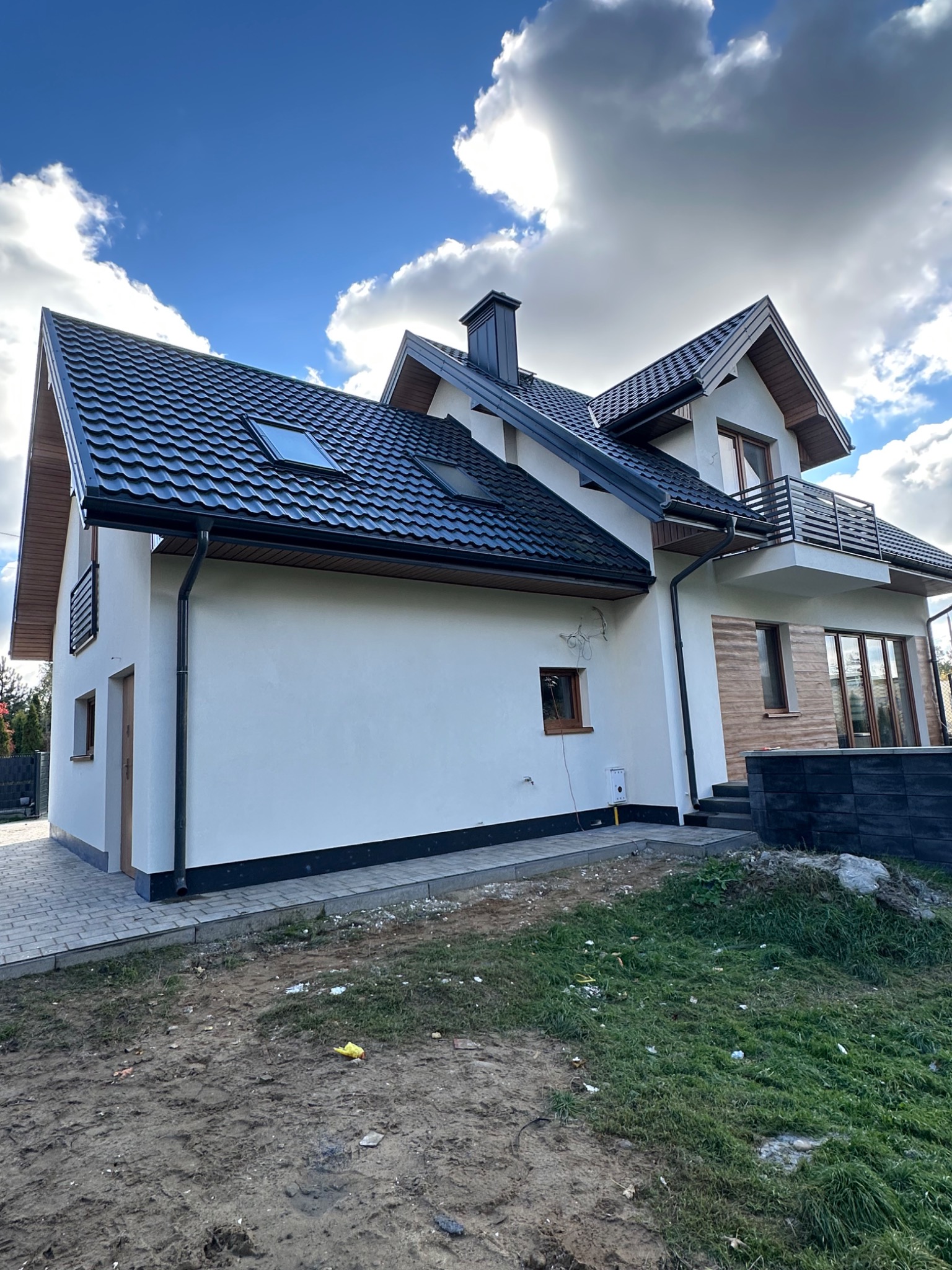 Dwukondygnacyjny dom z balkonem i czarnym dachem, wykończony jasnym tynkiem. Widoczne okna dachowe i elementy drewniane. Ujęcie z poziomu gruntu, błękitne niebo z chmurami w tle.