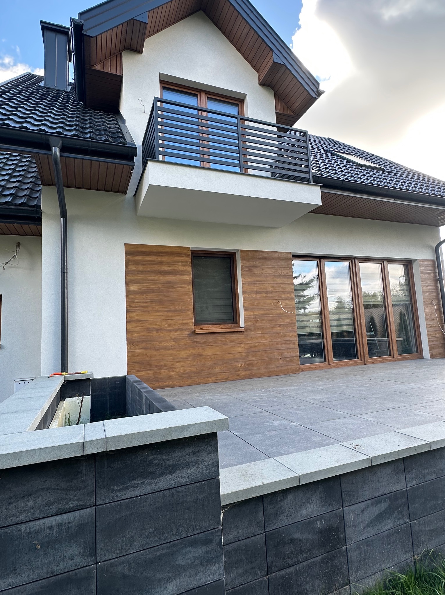 Nowoczesny dom jednorodzinny z balkonem i drewnianą elewacją, widok z poziomu tarasu. Eleganckie wykończenie, czarna dachówka i grafitowa kostka brukowa.