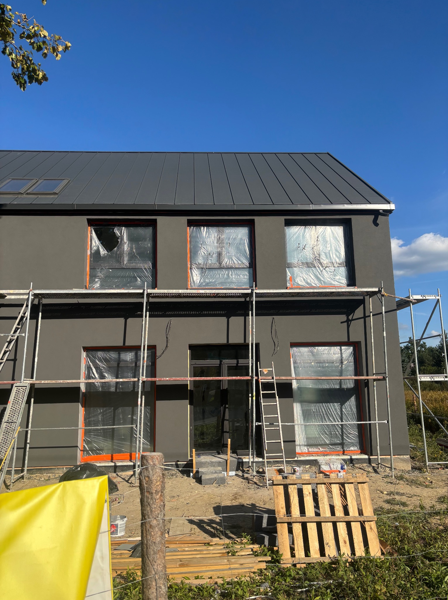 Dom w trakcie malowania elewacji na szaro, okna zabezpieczone folią, rusztowania, paleta drewniana na pierwszym planie, błękitne niebo w tle.