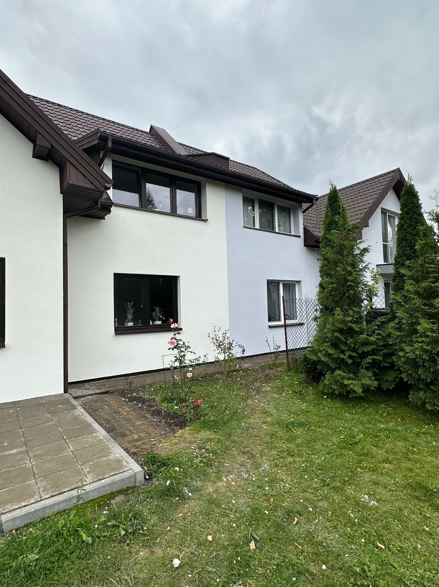 Dwukondygnacyjny dom z jasną elewacją, brązowym dachem i oknami. Część elewacji w odcieniu szarości. W tle pochmurne niebo i zieleń. Widok z poziomu gruntu.