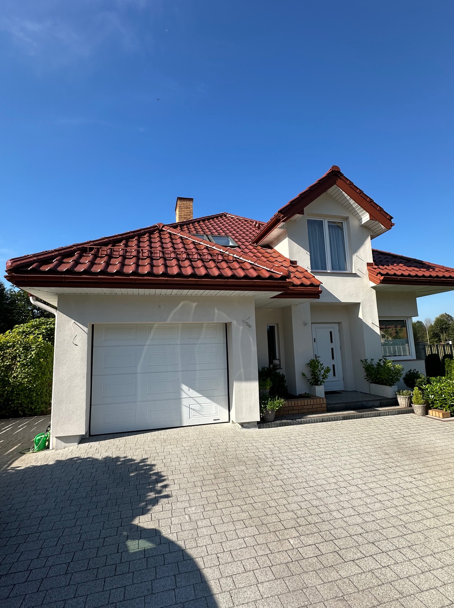 Biały dom jednorodzinny z czerwoną dachówką, garażem i brukowanym podjazdem w słoneczny dzień. Widok z poziomu gruntu, podkreślający estetykę elewacji.