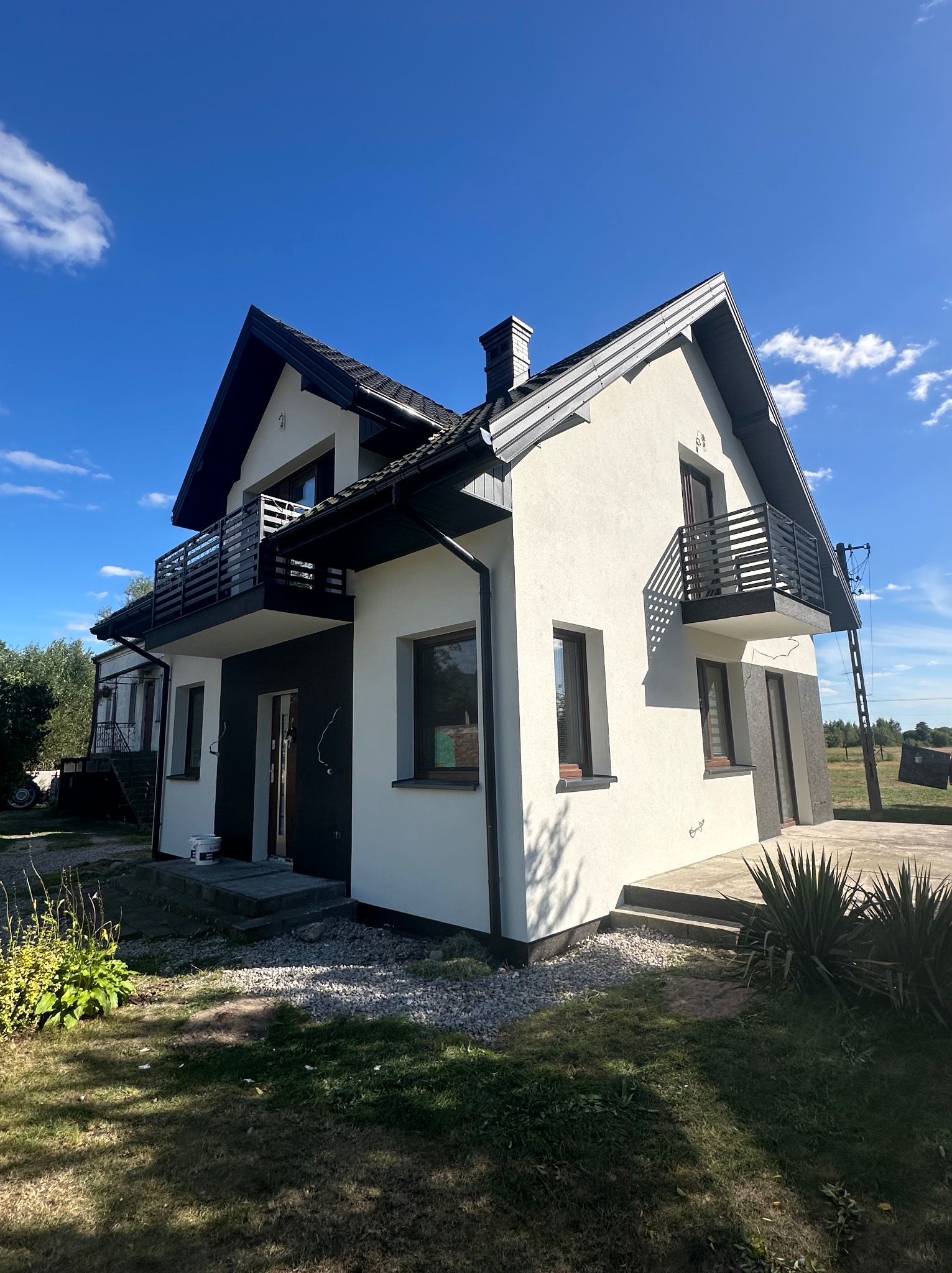 Dwukondygnacyjny dom z balkonami, wykończony w bieli i szarości, na tle błękitnego nieba. Widoczne elementy architektury i otoczenie domu.