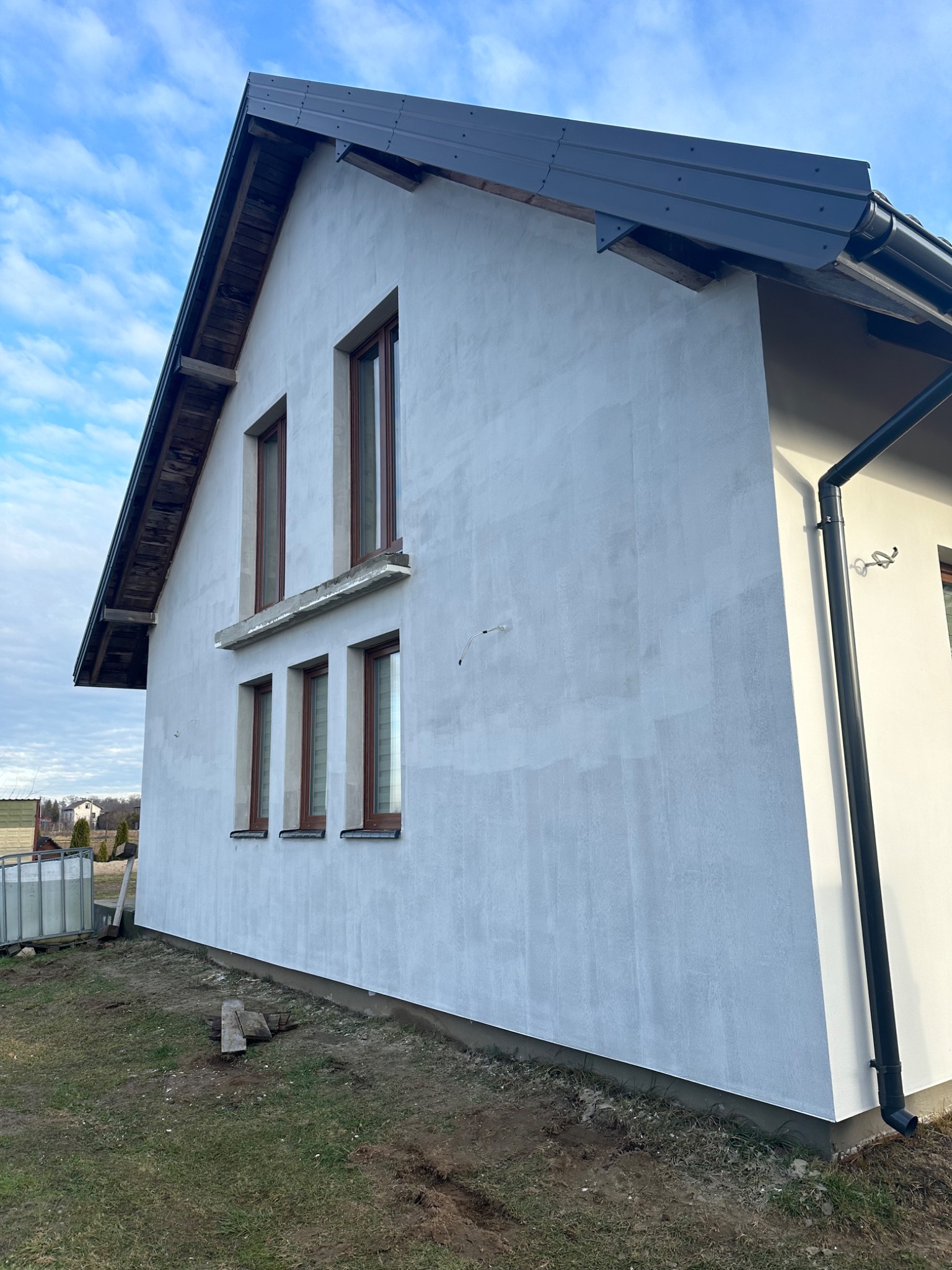 Elewacja domu jednorodzinnego po tynkowaniu, widoczne okna z brązowymi ramami i szara rynna, fragment dachu z metalowym pokryciem, trawa i częściowo odsłonięta ziemia wokół budynku.